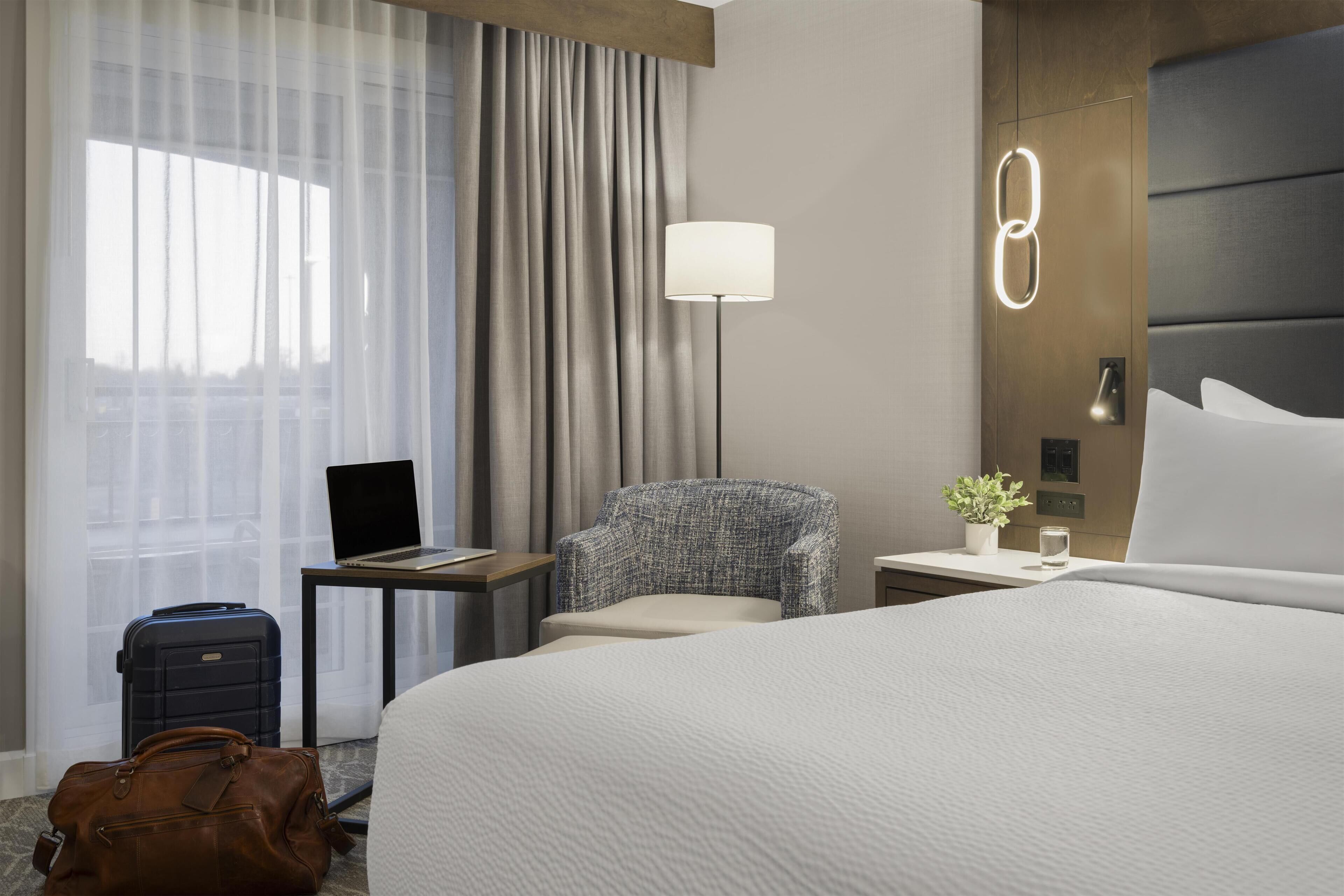 A hotel room with a bed, a desk with a laptop, a chair, a lamp, a suitcase, and a handbag.