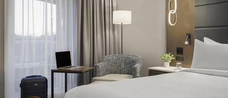 A hotel room with a bed, a desk with a laptop, a chair, a lamp, a suitcase, and a handbag.