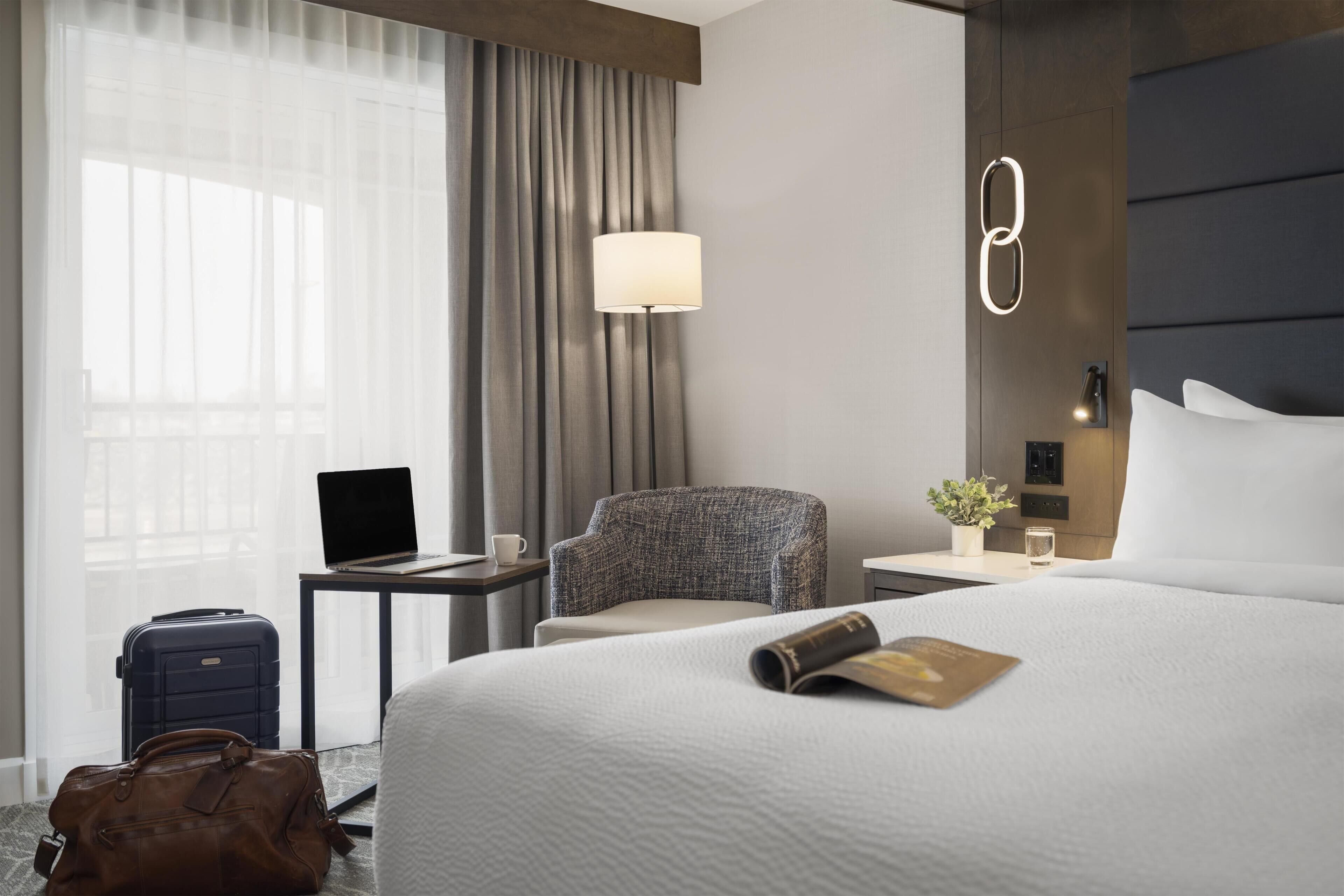 A hotel room with a bed, a desk, a chair, a lamp, a laptop, a suitcase, and a handbag.