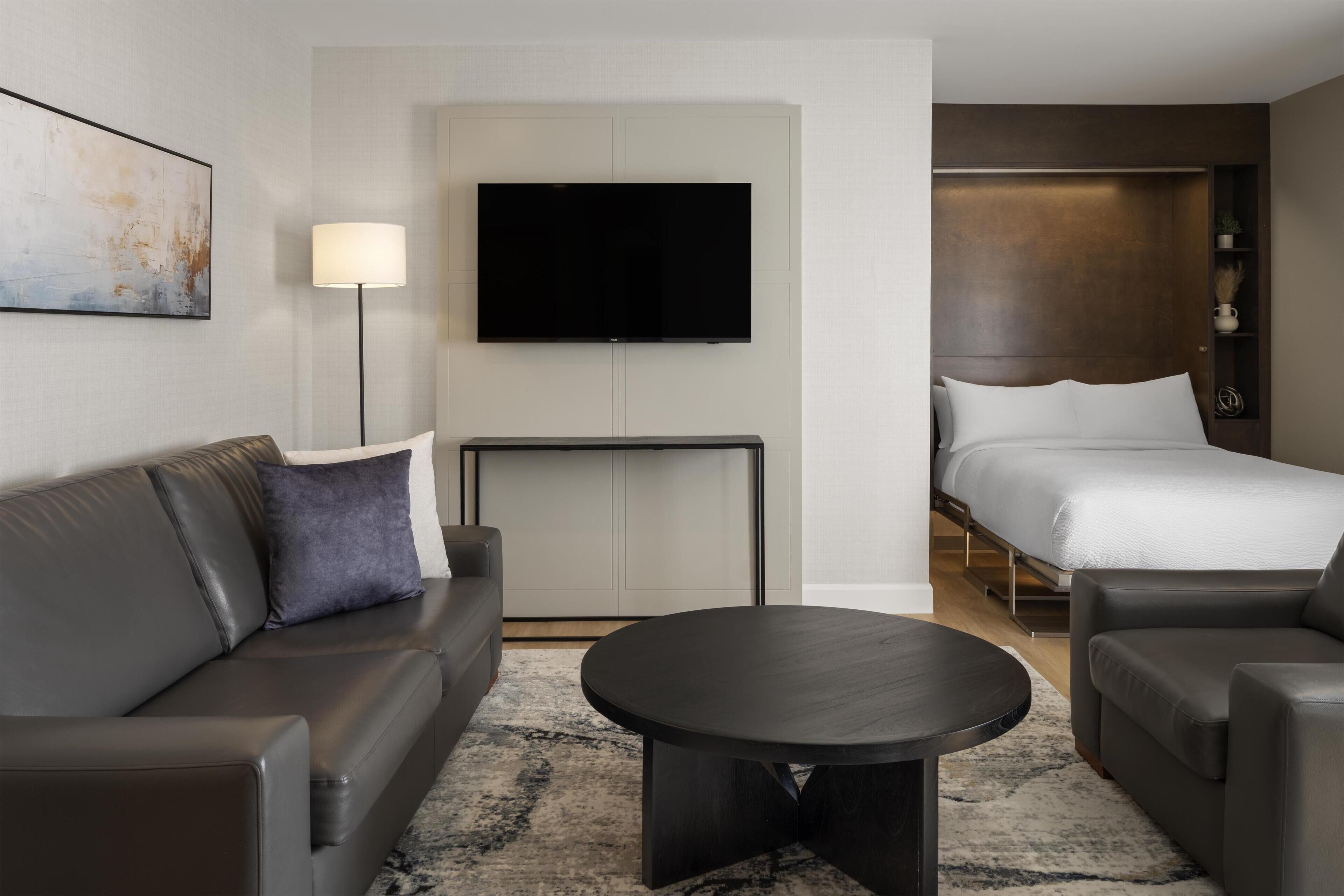 A modern hotel room with a sofa, a round coffee table, a flat-screen TV, a bed, and a nightstand.
