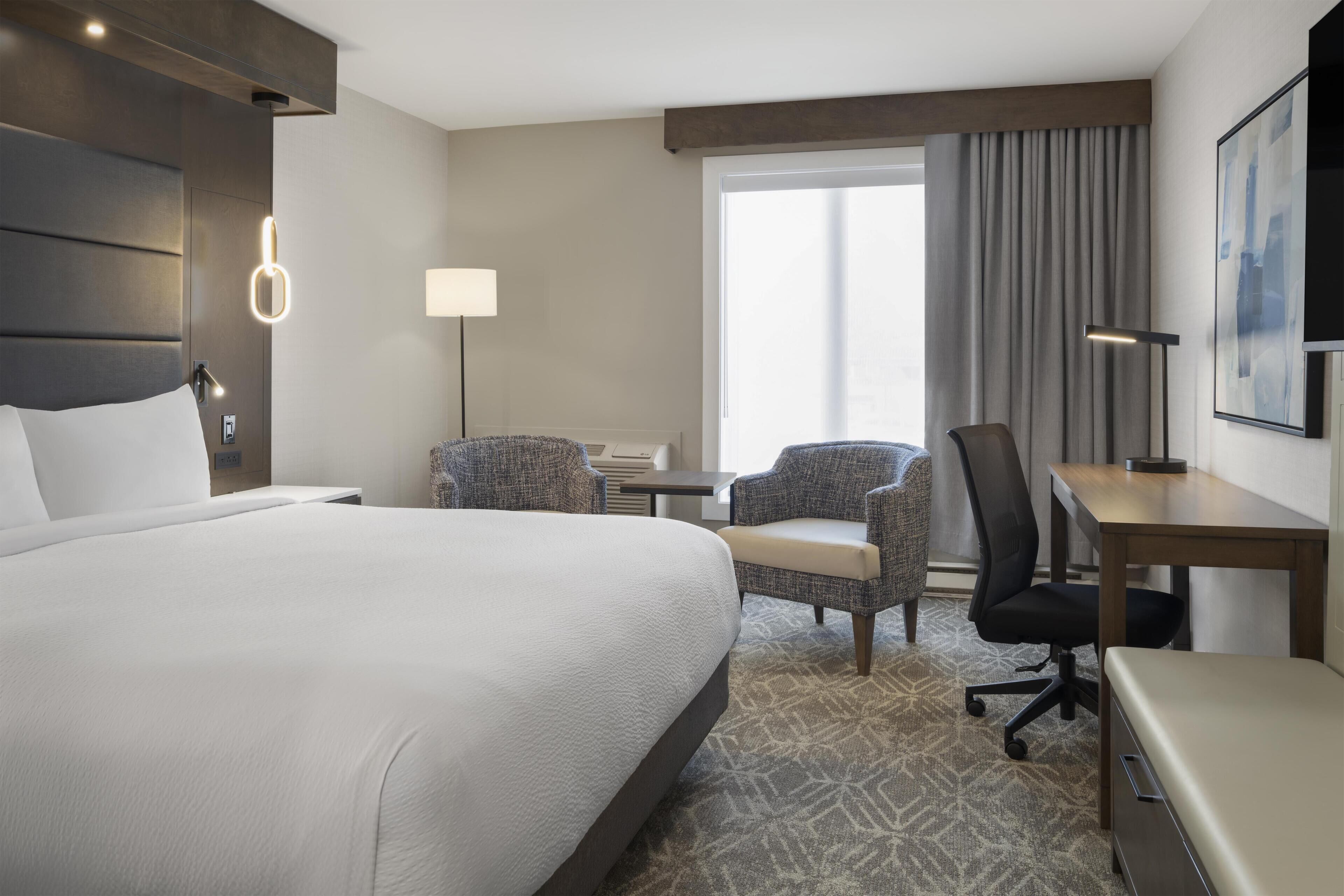 A hotel room with a large bed, two armchairs, a desk, a chair, a lamp, and a large window with curtains.