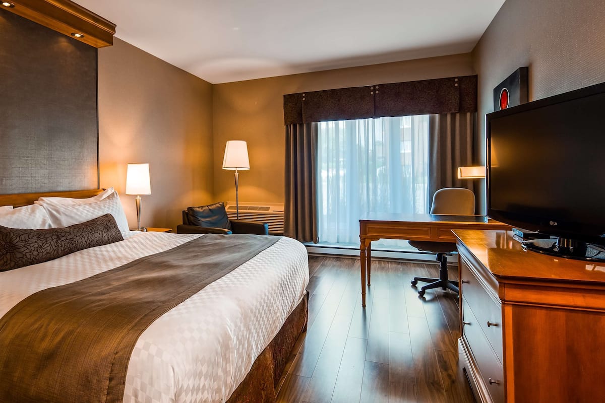 A hotel room with a large bed, a desk with a flat-screen TV, a chair, a lamp, and a window with curtains.