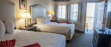 Quarto de hotel com duas camas, uma escrivaninha com luminária, cômoda e vista para a varanda.