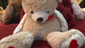 Un oso de peluche suave con un lazo rojo y patas en forma de corazón sentado sobre una superficie tapizada en rojo.