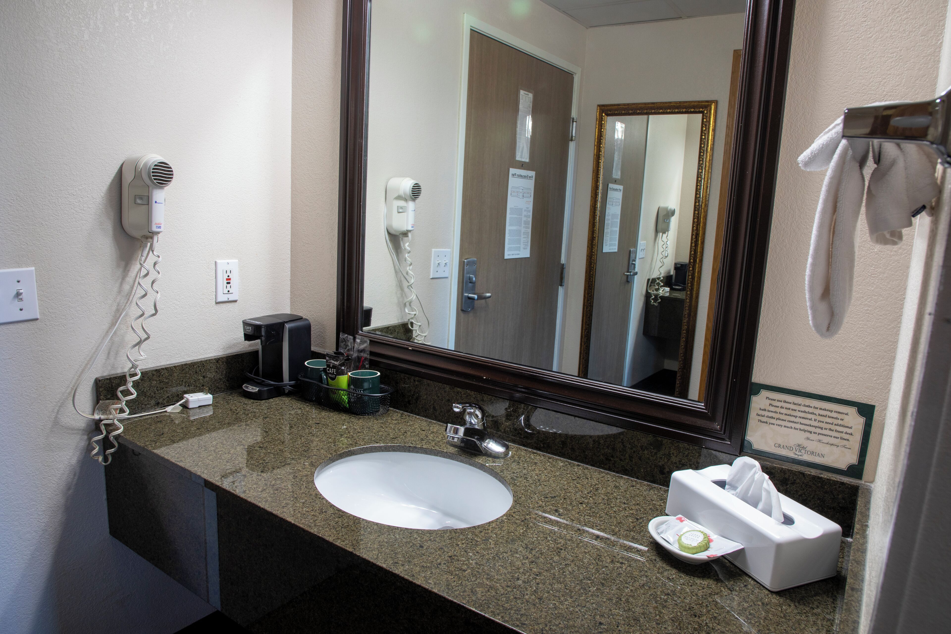 Baño de hotel con encimera de granito, lavamanos blanco, secador de pelo, espejo y toallero.