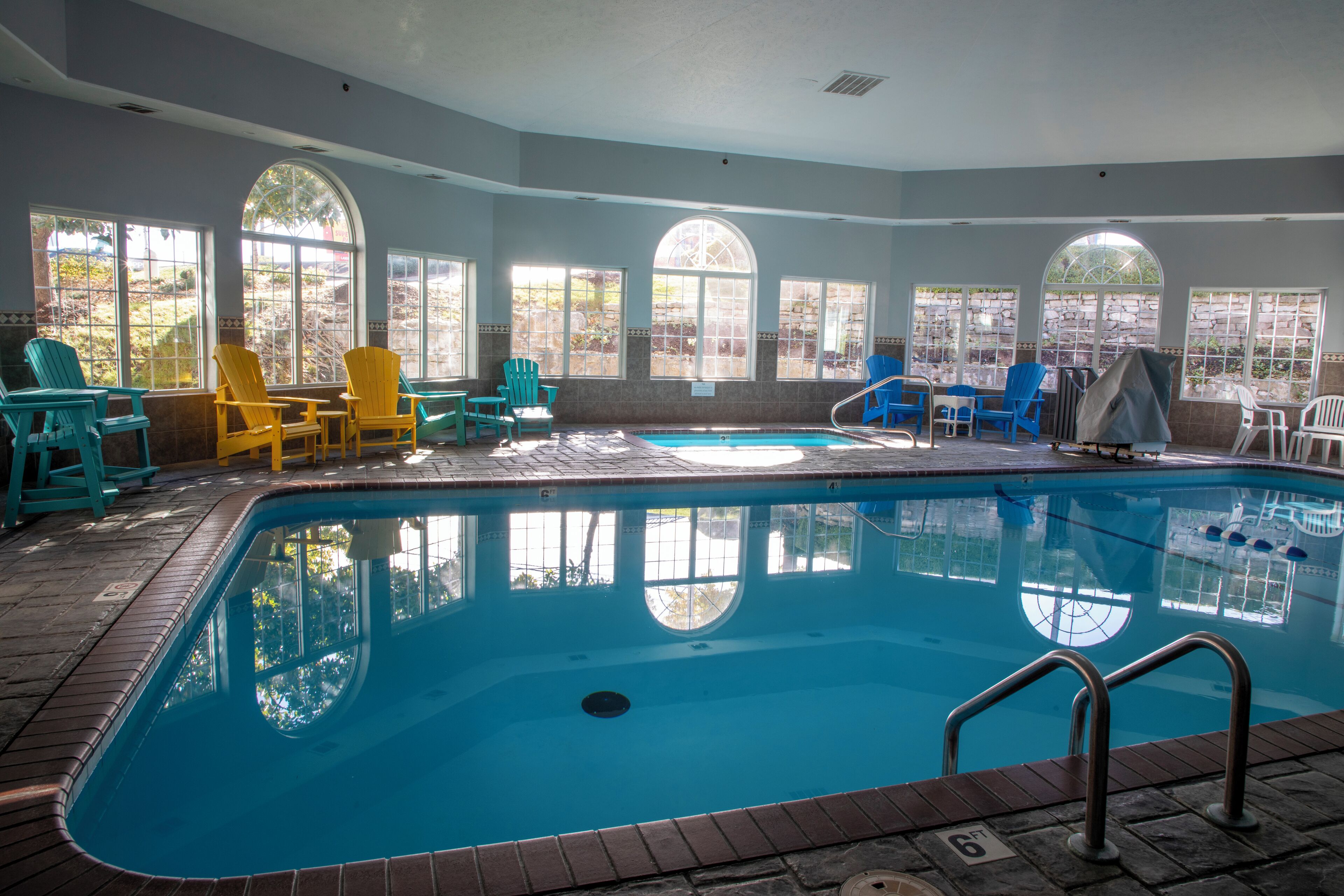 An indoor swimming pool with clear views through large windows, surrounded by various chairs and tables.