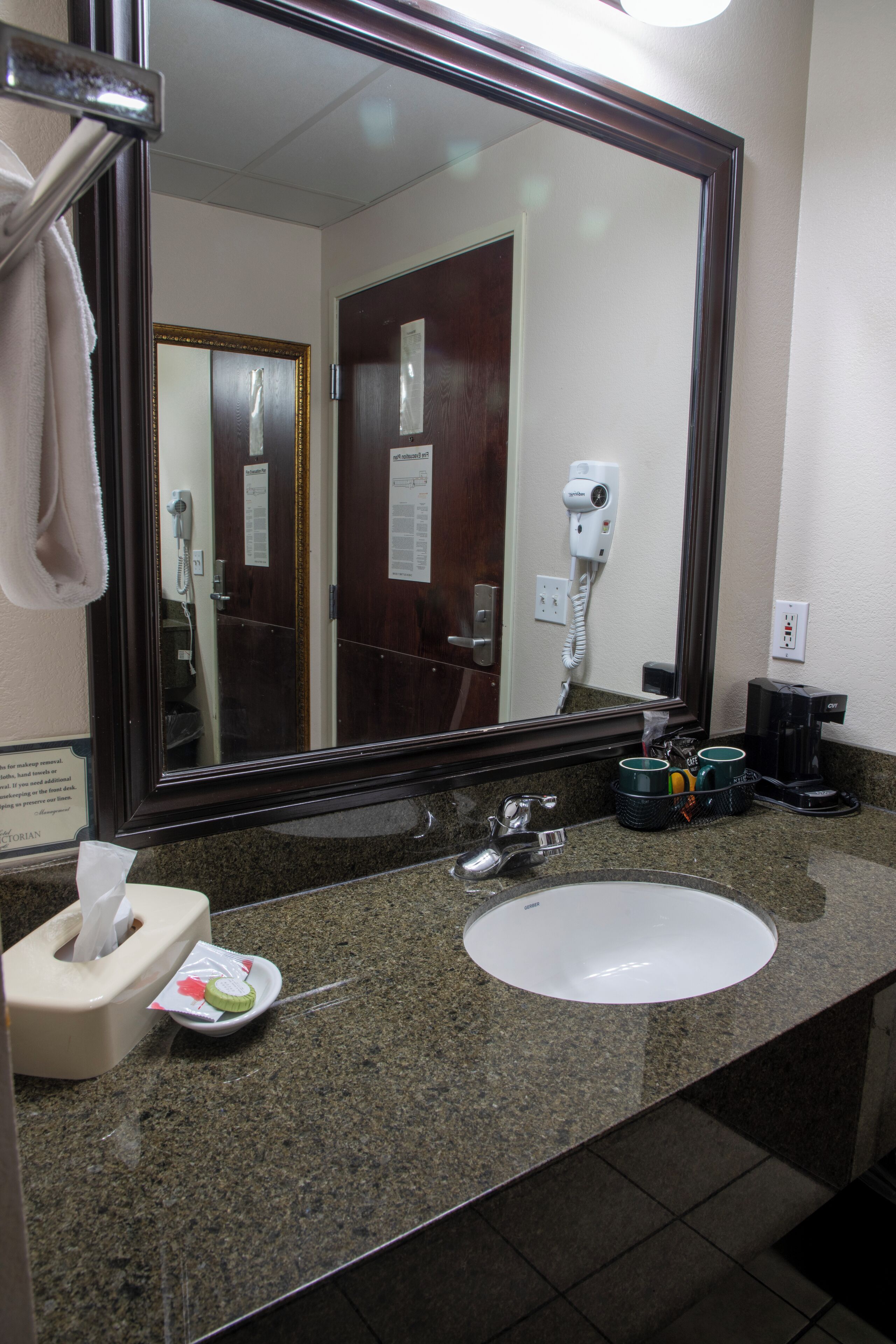 Un baño de hotel con un espejo grande, un lavamanos, dispensador de toallas, secador de pelo y cafetera.