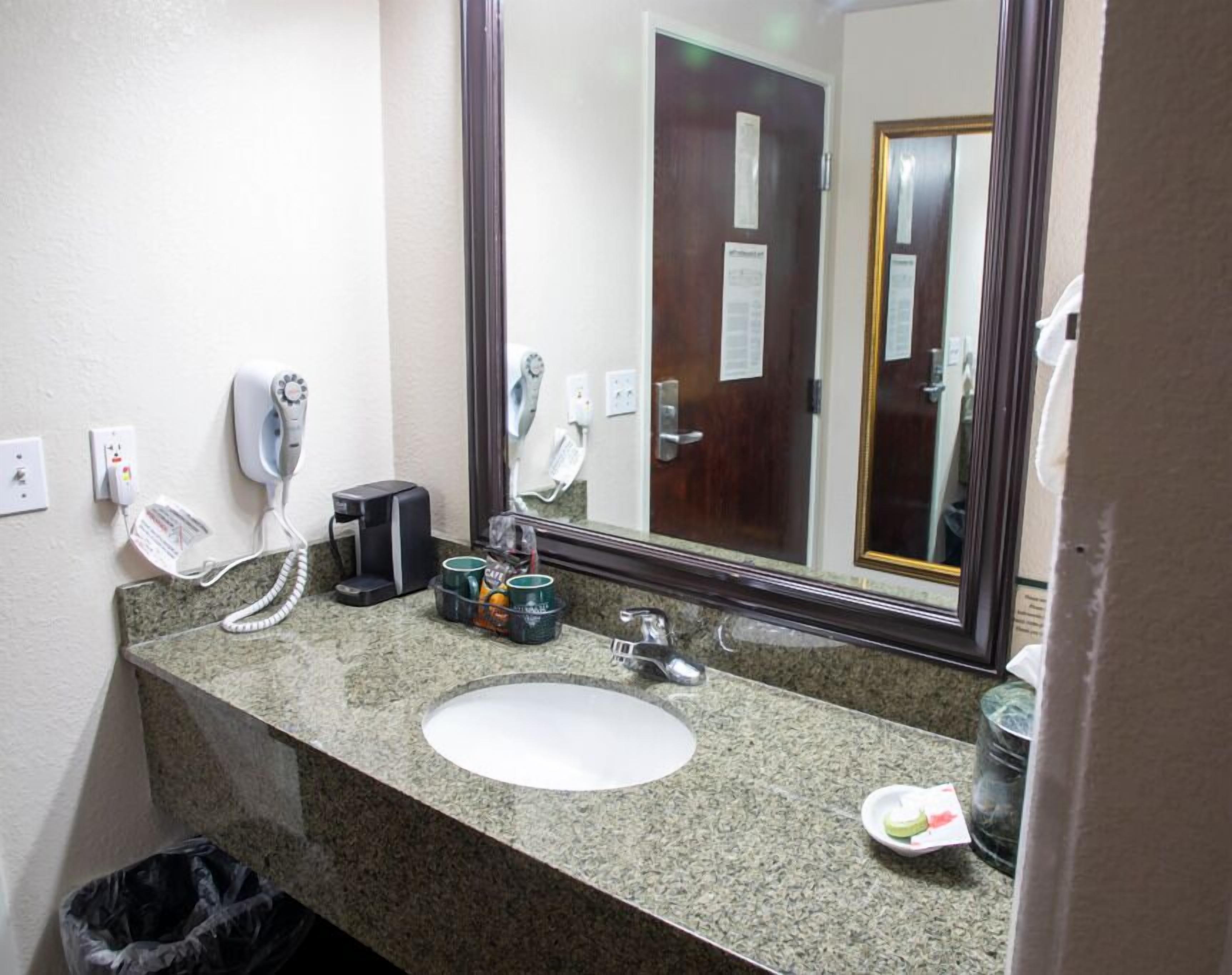 Ein Hotelbad mit einer Granit-Arbeitsplatte, einem weißen Waschbecken, einer Kaffeemaschine, einem Föhn und einem Mülleimer.