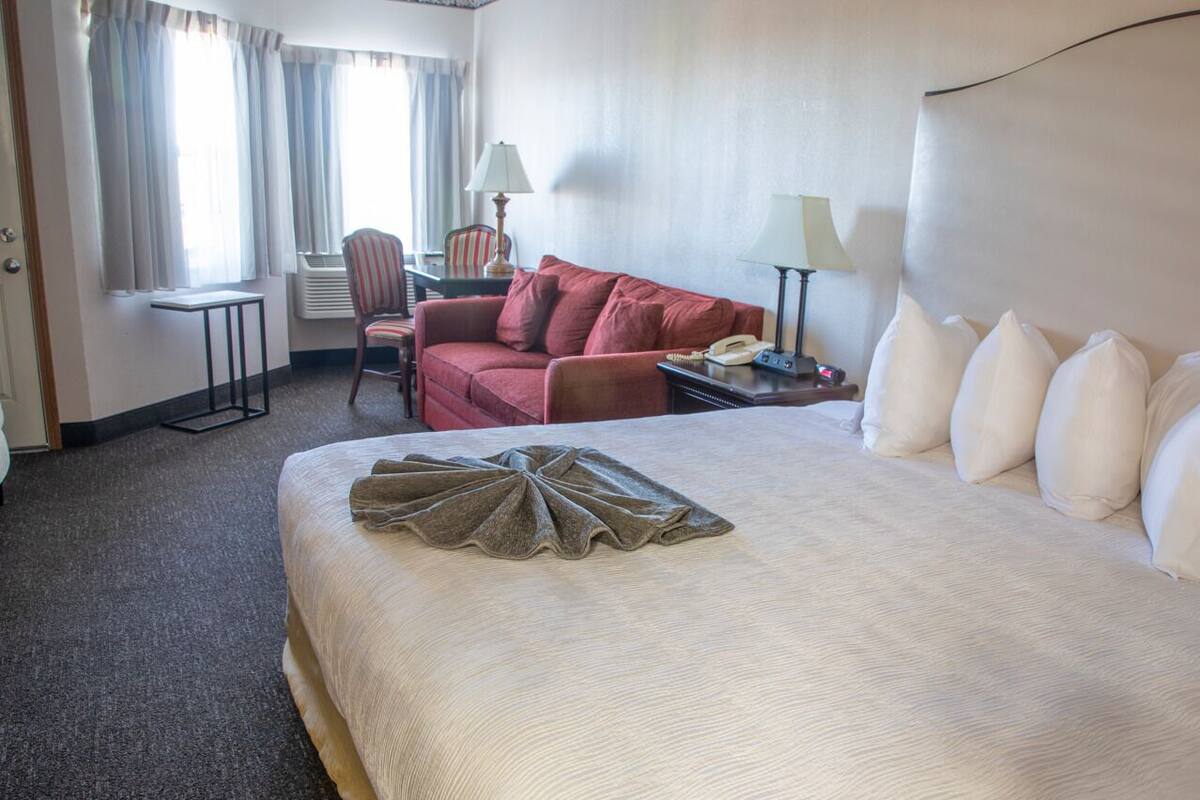 A hotel room with a large bed, a red sofa, a small table with a lamp, and a chair.