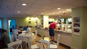 A restaurant with round tables and white chairs, a counter with a chef preparing food, and a green wall with shelves.