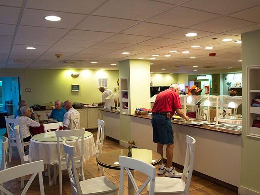 A restaurant with round tables and white chairs, a counter with a chef preparing food, and a green wall with shelves.