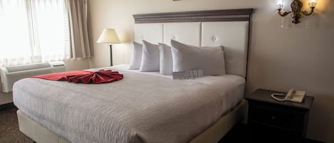 Une chambre d’hôtel avec un grand lit, une table de chevet avec un téléphone, une commode, une lampe et une fenêtre avec des rideaux.