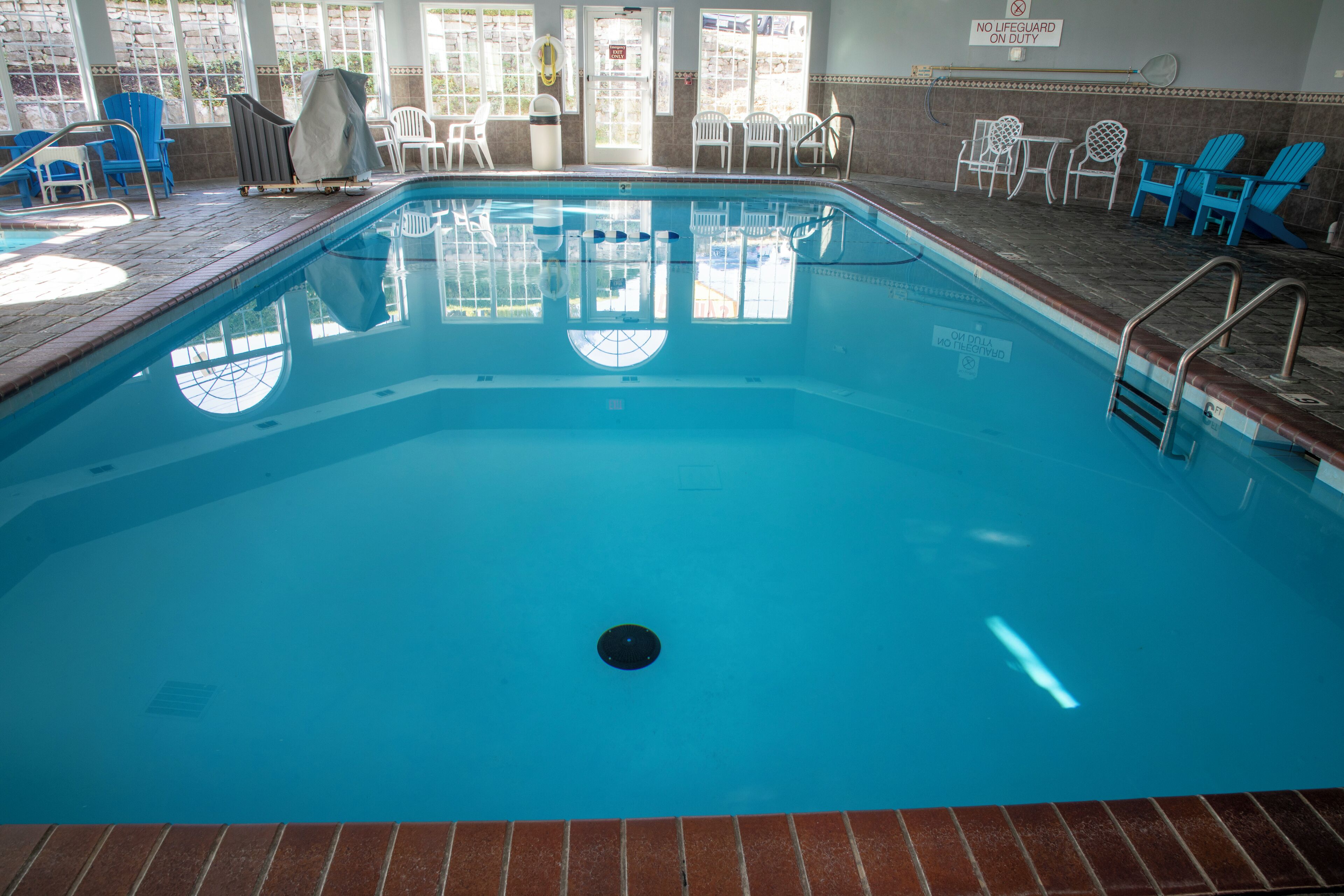 An indoor swimming pool with clear blue water, surrounded by blue and white chairs, and a sign indicating no lifeguard on duty.