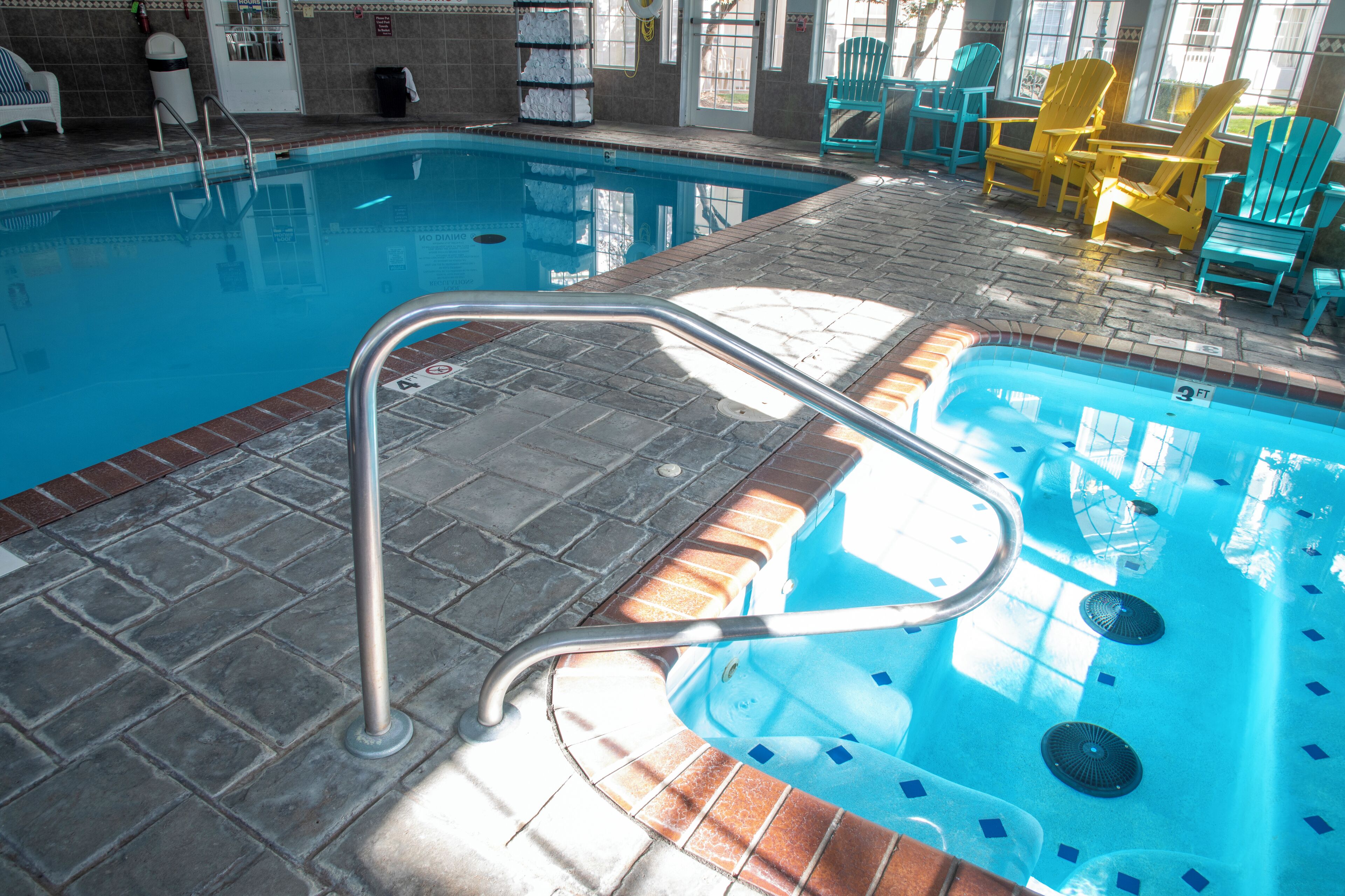 Una piscina con una escalera metálica, suelo de azulejos y varias sillas alrededor de la zona de la piscina.
