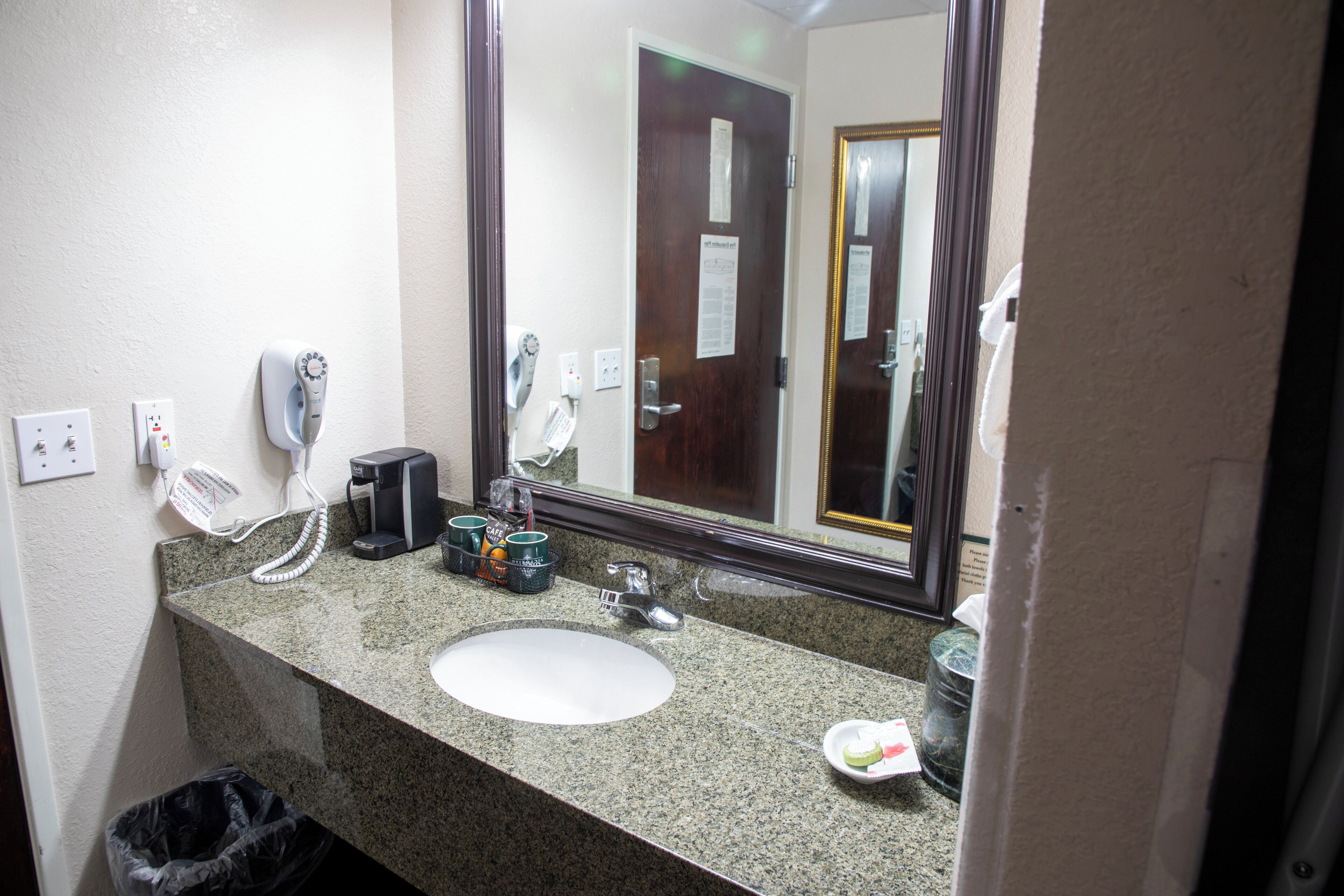 Une salle de bain dotée d’un comptoir en granit, d’un lavabo blanc, d’un sèche-cheveux, d’une machine à café et d’une poubelle.