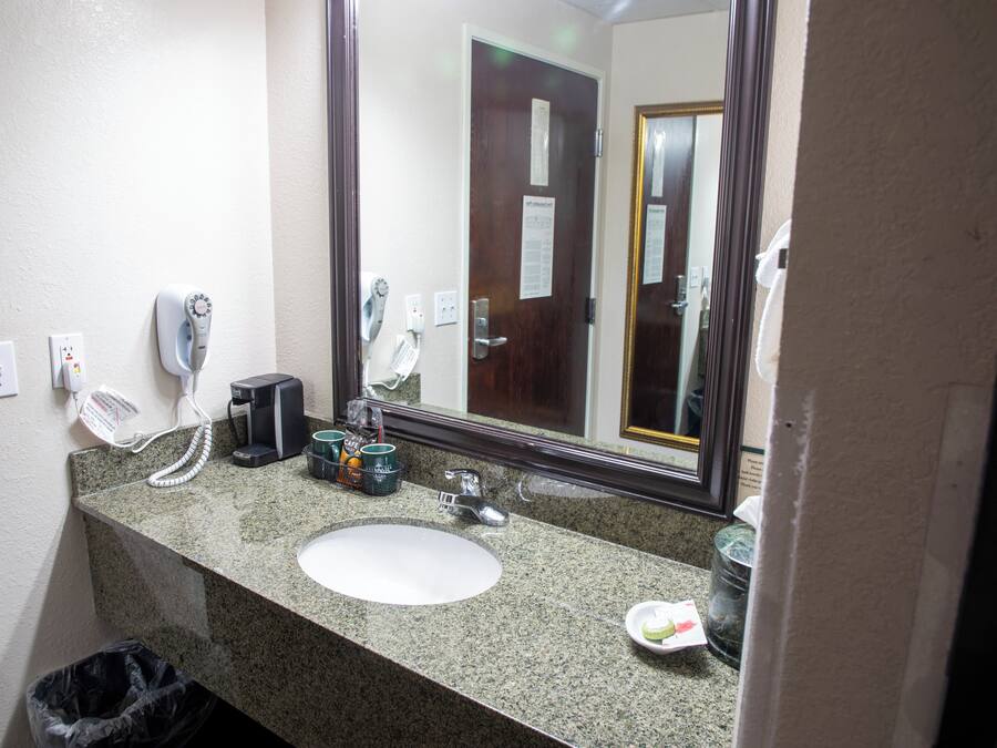 A bathroom with a granite countertop, a white sink, a hair dryer, a coffee maker, and a trash can.