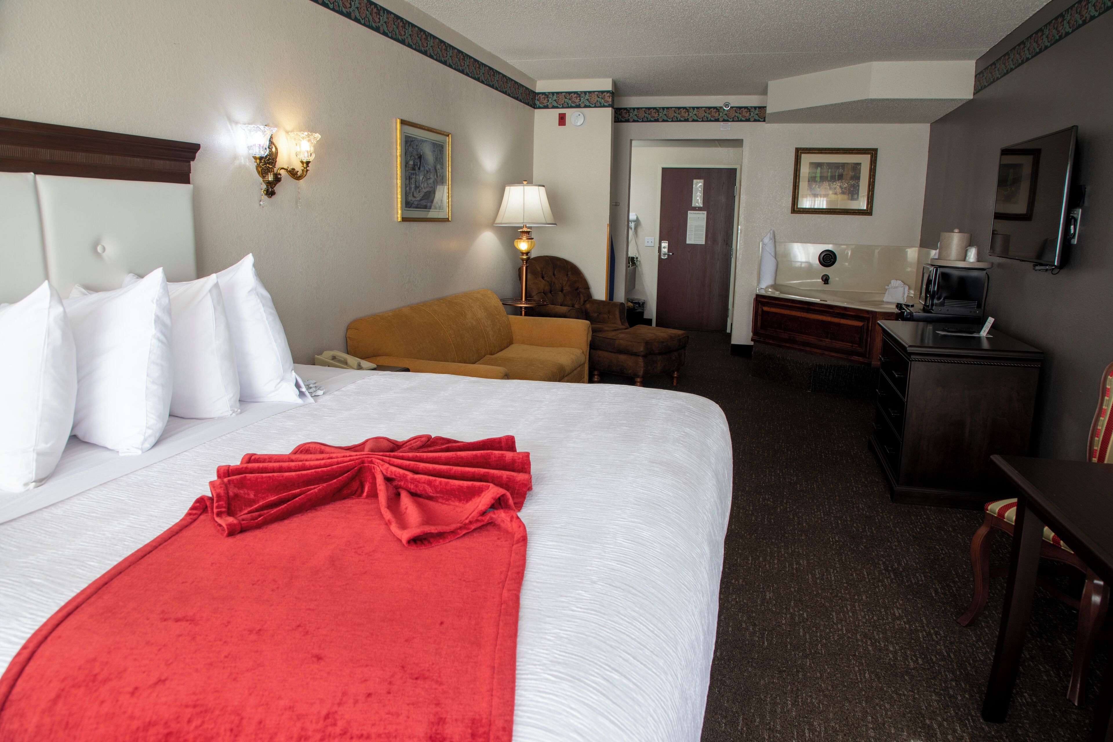 A hotel room with a large bed, a red blanket, a sofa, a desk with a chair, and a television.