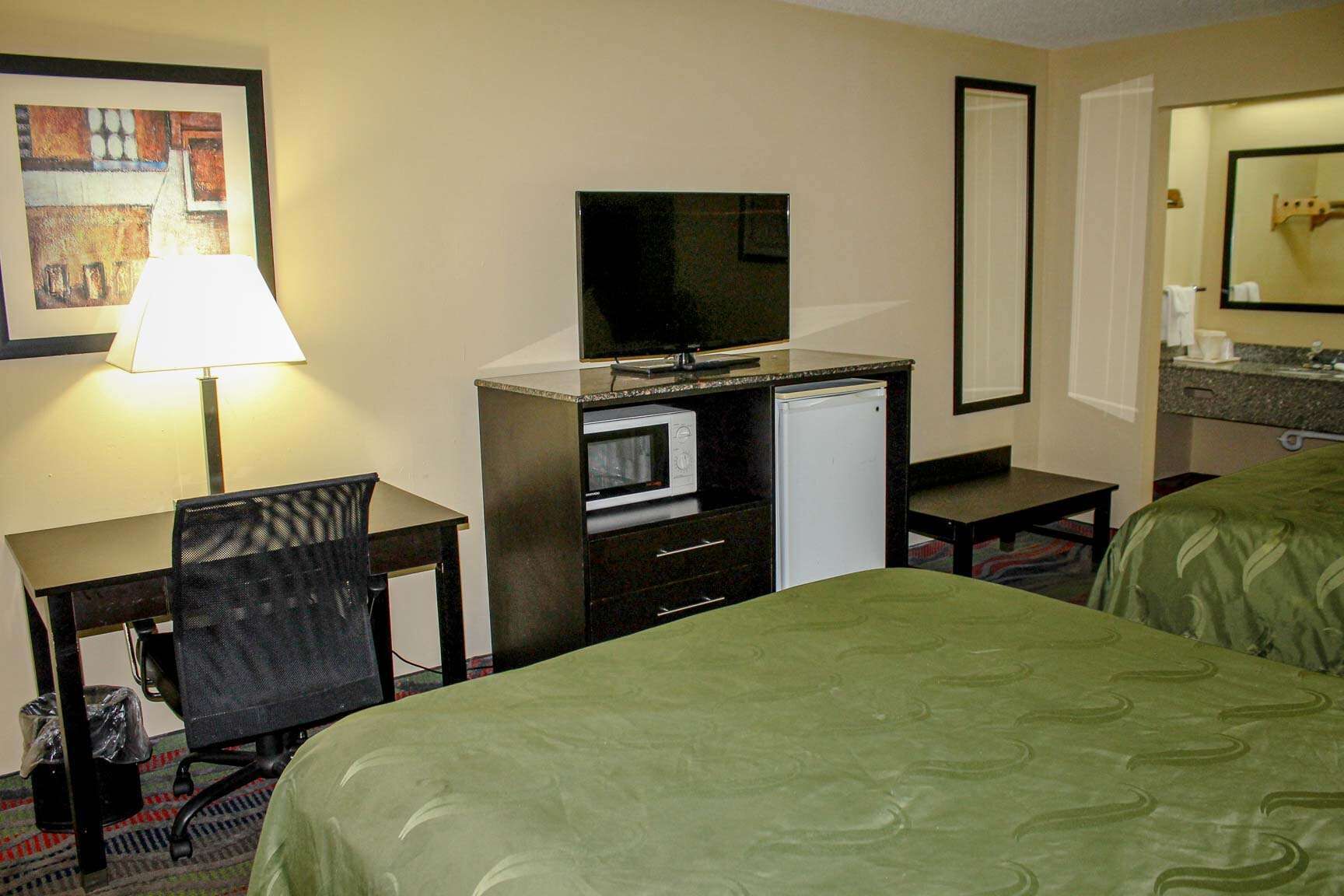 Une chambre d’hôtel comprenant un lit, un bureau avec une chaise, une télévision, un réfrigérateur et une salle de bain avec un miroir.