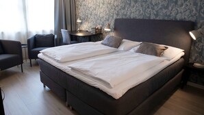 Una cama doble con sábanas blancas, un cabecero oscuro y dos lámparas de noche. Junto a la cama hay una pequeña mesa con una lámpara y algunos libros. La habitación tiene papel tapiz estampado y una ventana con cortinas.