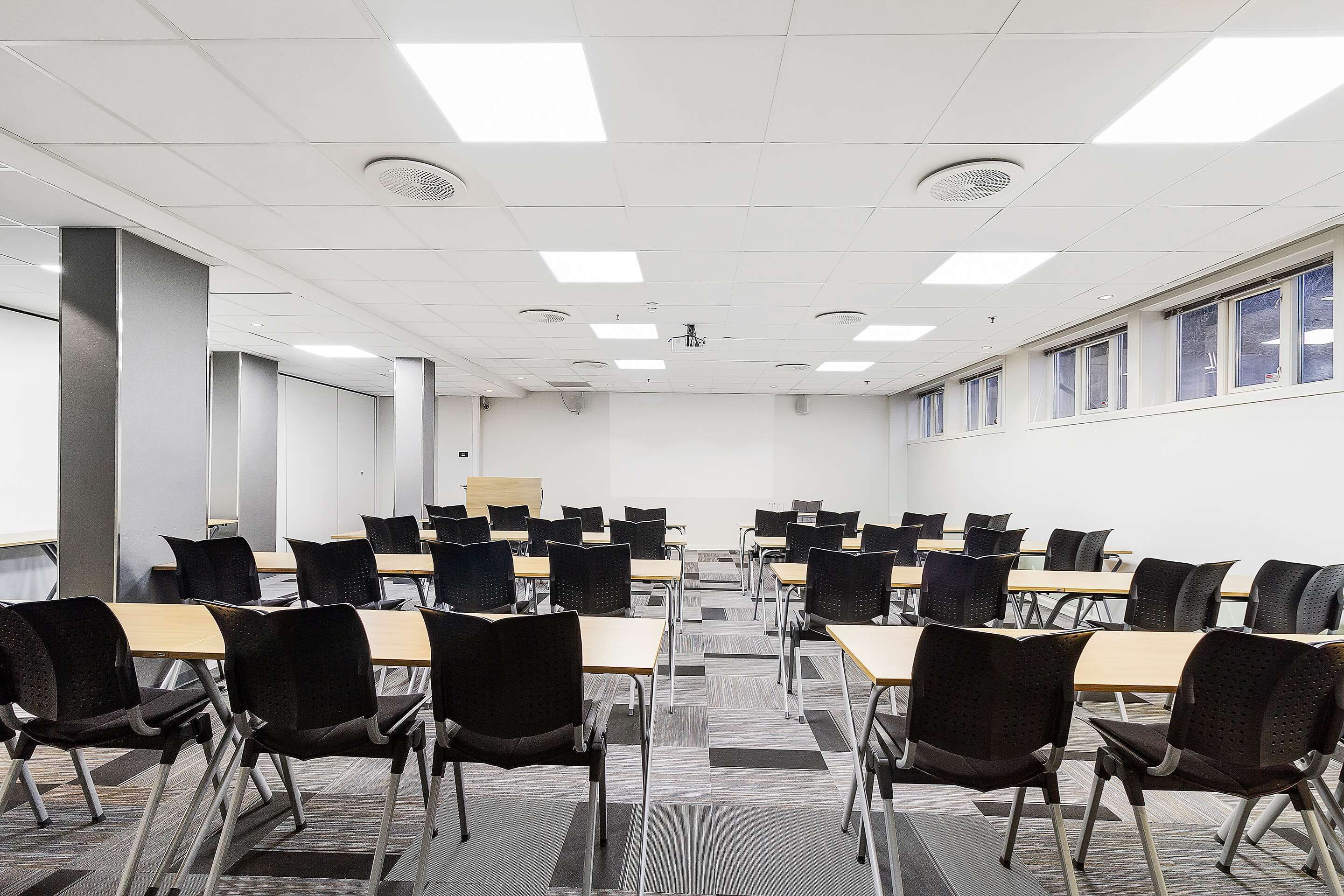 A conference room with rows of tables and chairs, white walls, and a projector screen.