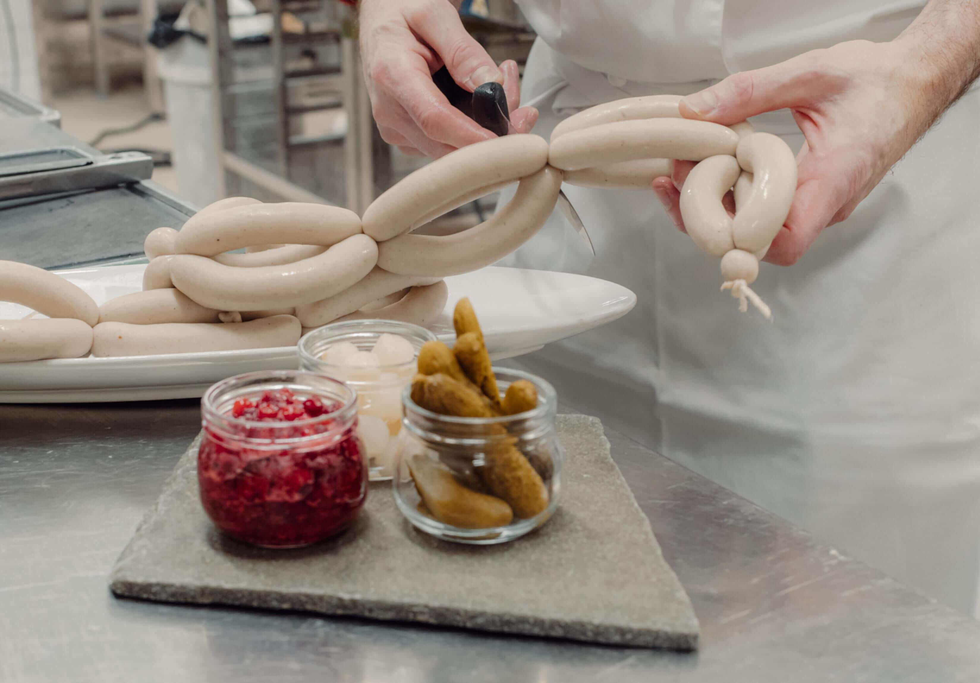 A person preparing sausages on a stainless steel counter with jars of spices and cashews.