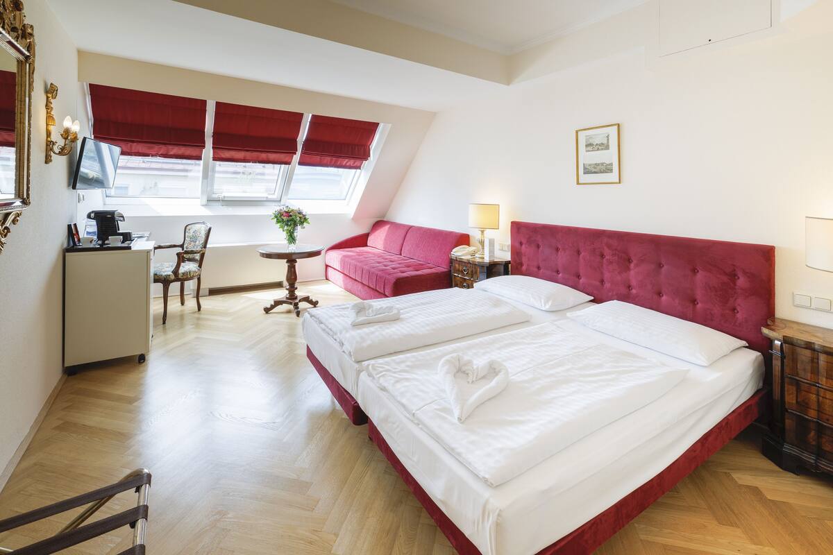 A hotel room with a large bed, a desk with a computer, a chair, a small table with a lamp, and a window with red blinds.