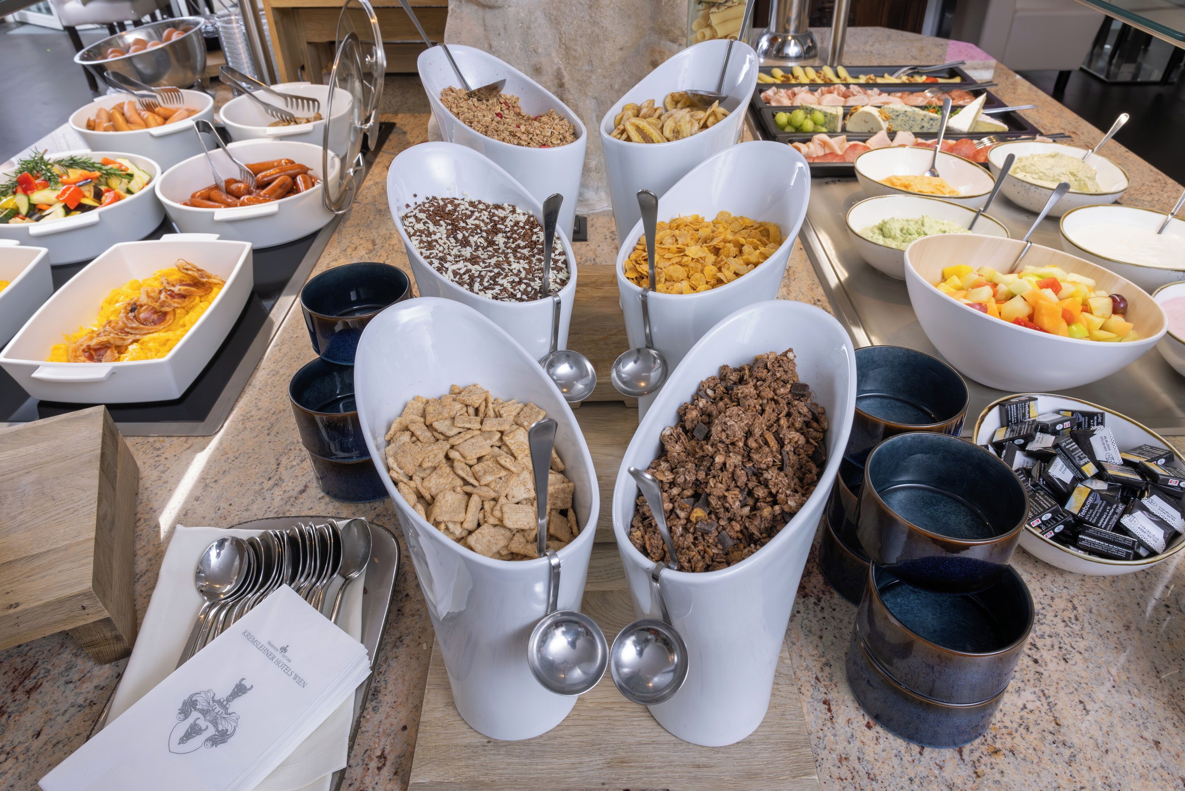 A buffet with various food items in white bowls and trays, including fruits, vegetables, and cereals.