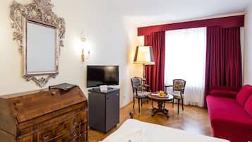Una habitación de hotel con un aparador de madera, un televisor de pantalla plana, un escritorio con lámpara, un sofá rojo y un ventanal con cortinas rojas.