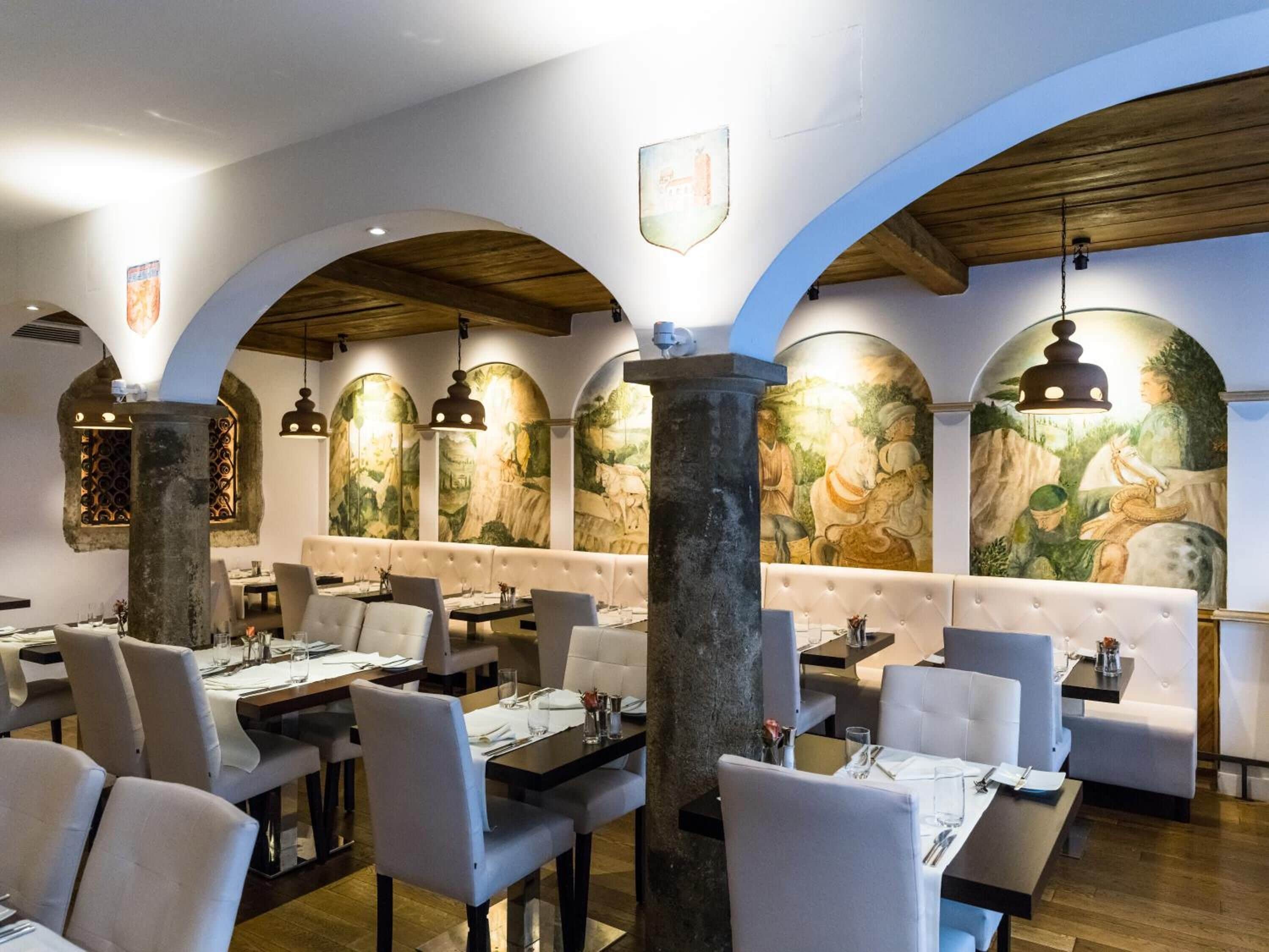 Restaurante com altos tetos arqueados, colunas de pedra e pinturas murais. Mesas montadas com toalhas brancas e cadeiras.
