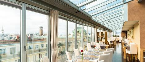 A restaurant with long tables set for dining, large windows offering city views, and a modern interior design.