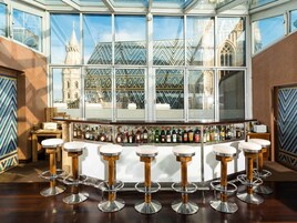 Un área de bar con barra alta y varios taburetes, amplias ventanas con vista a un edificio histórico y una variedad de botellas sobre la barra.