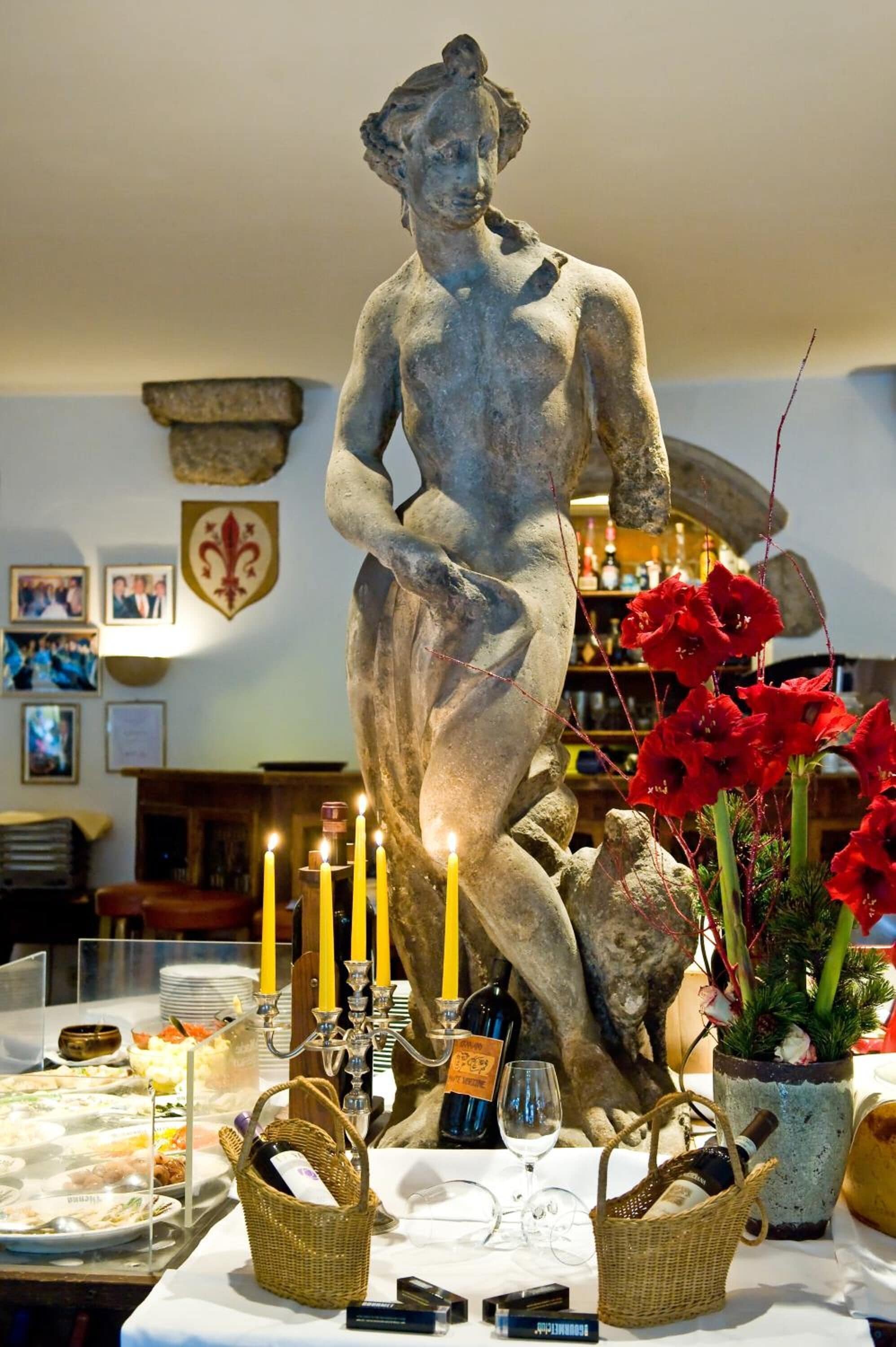 A statue of a woman in a room with a table set for a meal, candles, and a wine bottle.