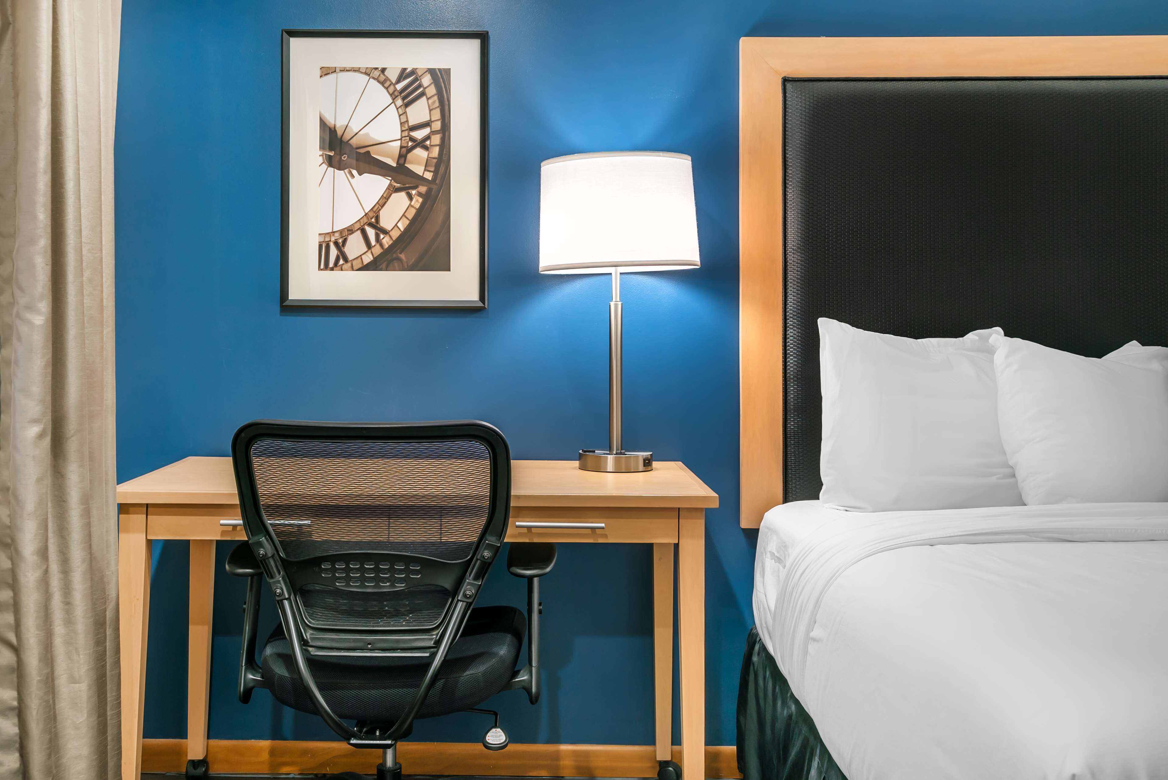 A hotel room with a bed, a desk with a chair, a lamp, and a framed picture on the wall.