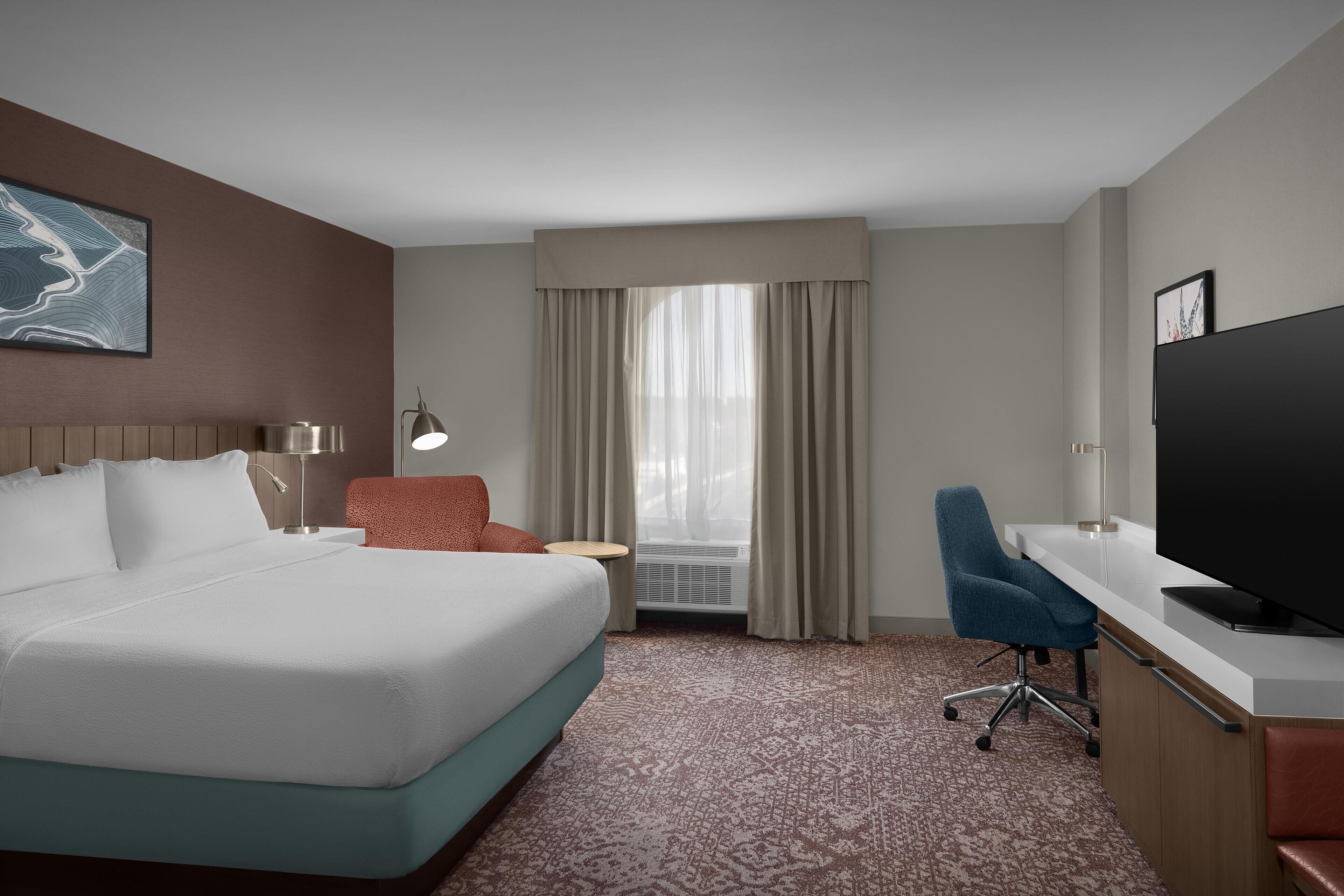 A hotel room with a large bed, a desk with a television, a chair, a small table with a lamp, and a window with curtains.