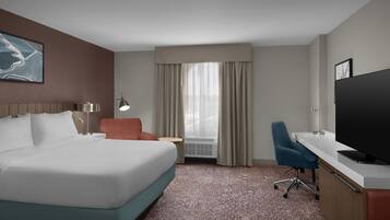 A hotel room with a large bed, a desk with a television, a chair, a small table with a lamp, and a window with curtains.