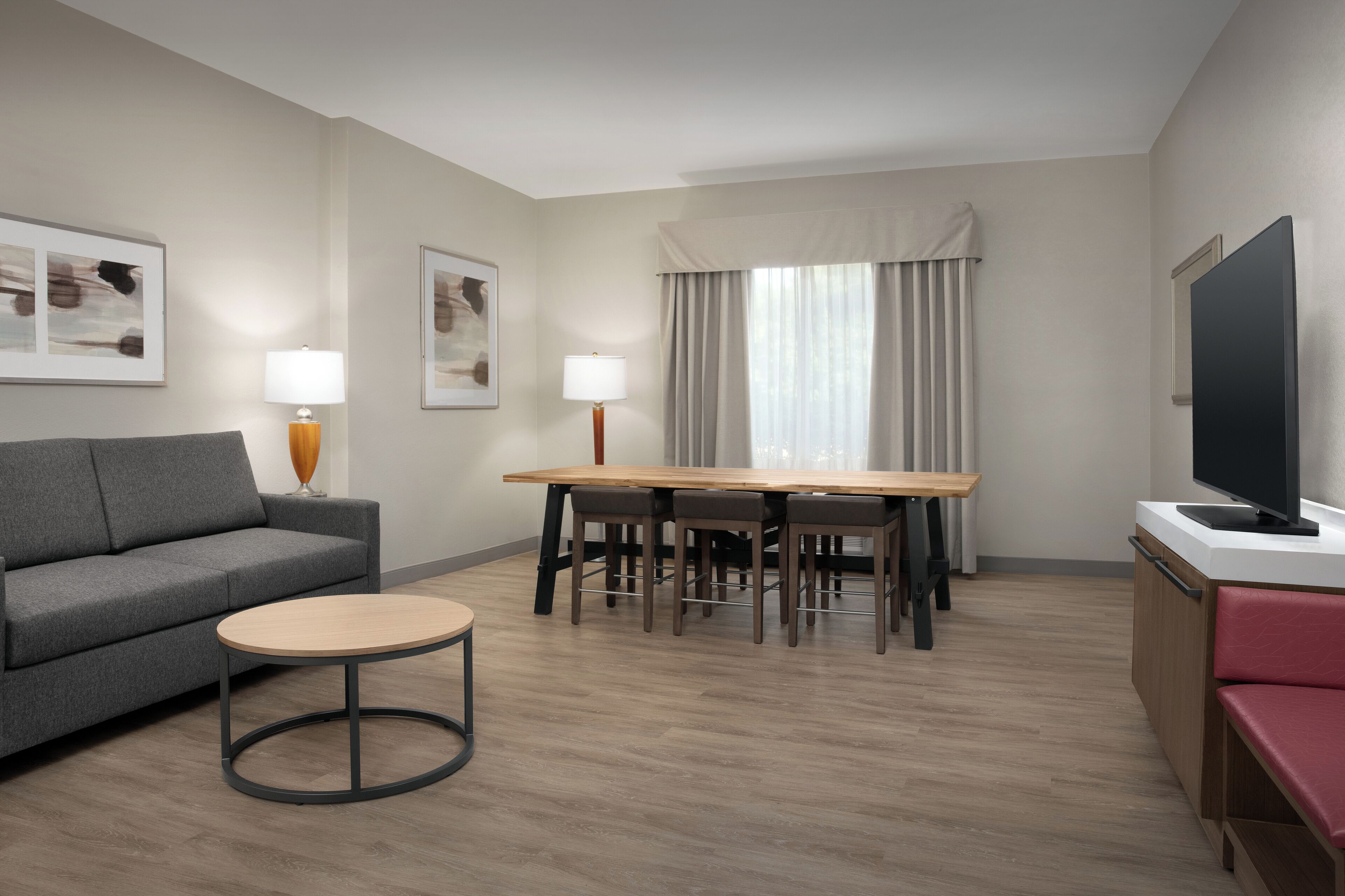 Una habitación de hotel moderna con un sofá gris, una mesa de centro redonda, una mesa de comedor de madera con sillas, un televisor y obras de arte en las paredes.