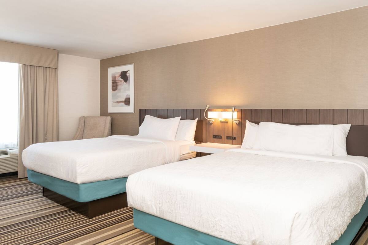 A hotel room with two beds, a headboard with integrated lighting, a framed picture on the wall, and a window with curtains.