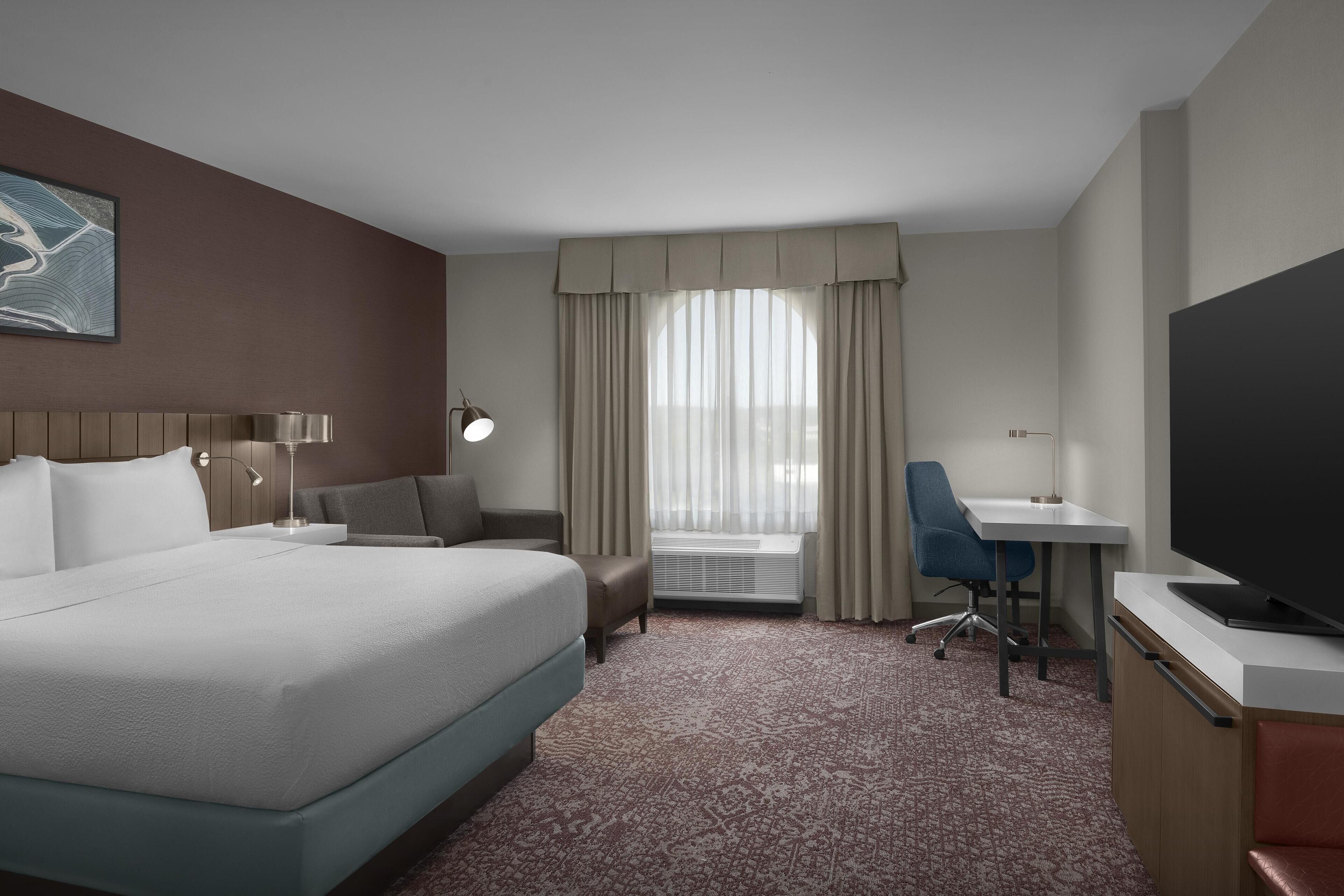 A hotel room with a large bed, a desk, a chair, a TV, and a window with curtains.