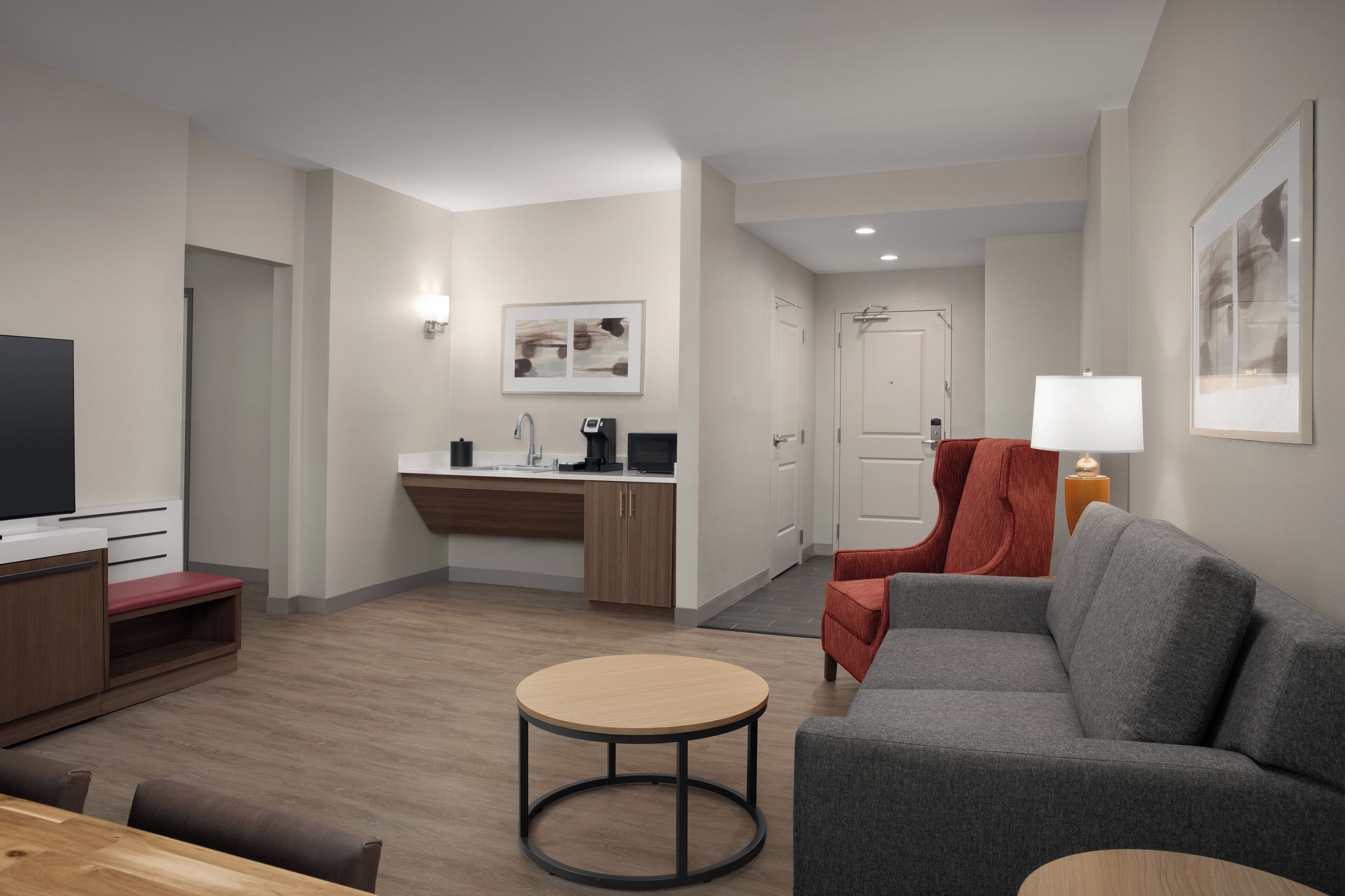 A modern living room with a grey sofa, a round coffee table, a TV, and a kitchen area with a sink and countertop.