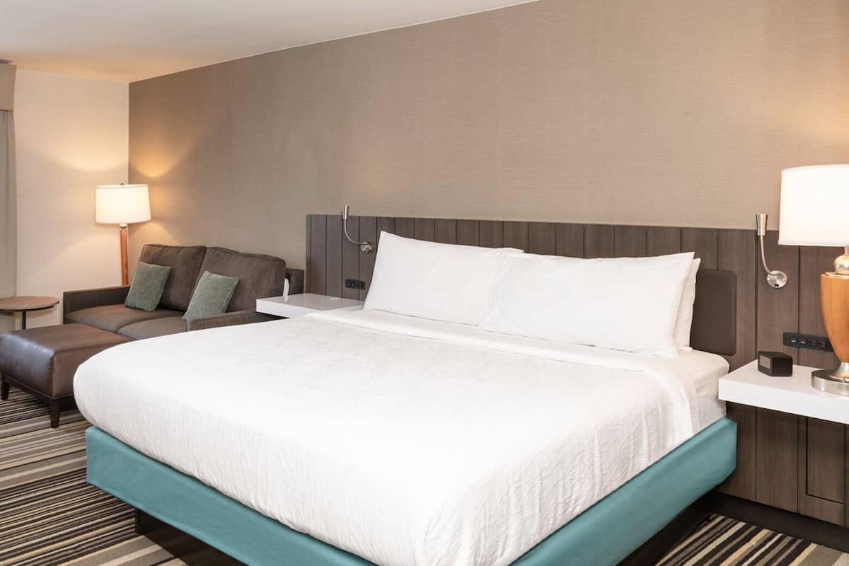 A hotel room with a large bed, a sofa, a side table, a lamp, and a window with curtains.