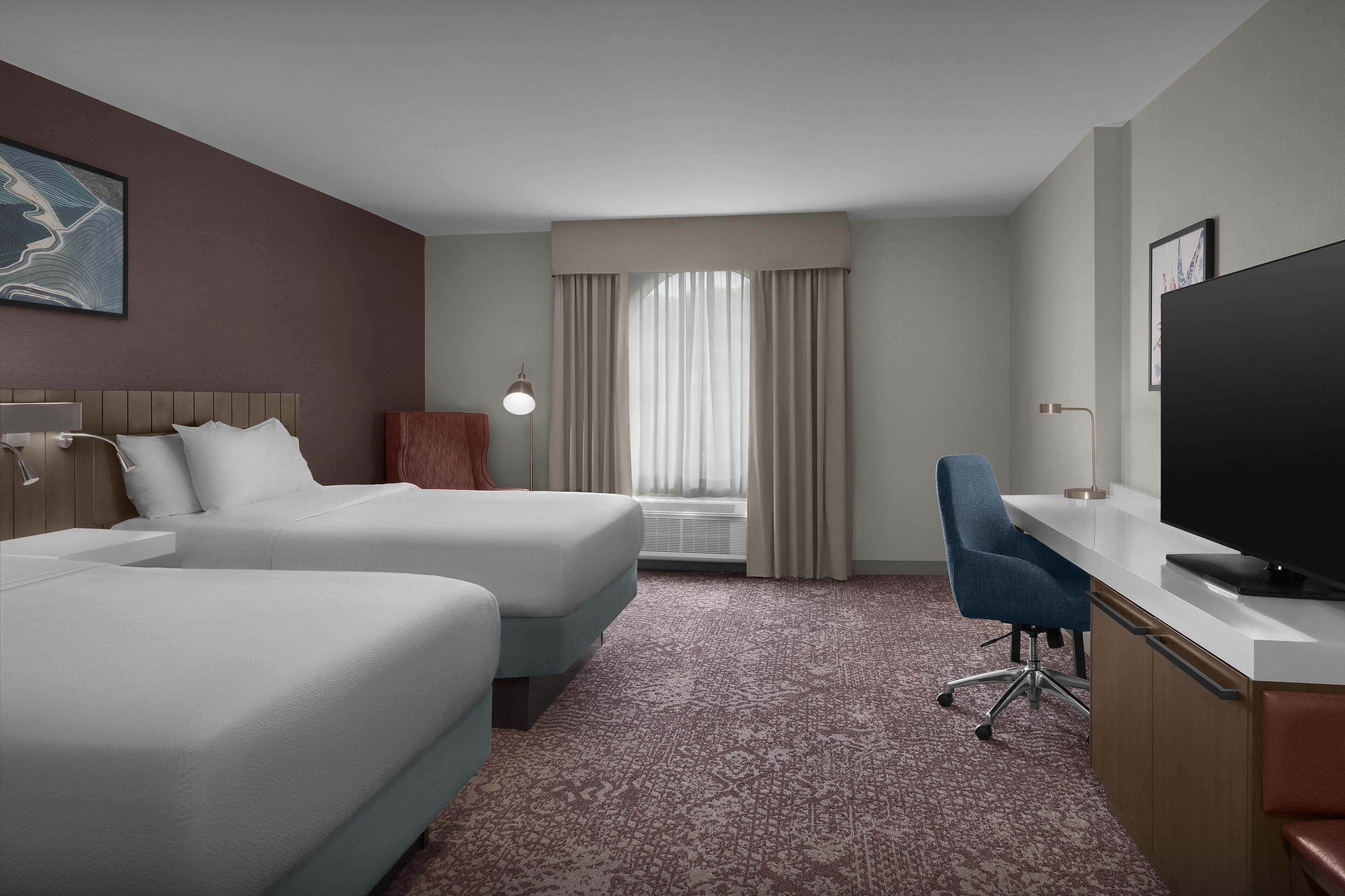 Quarto de hotel com duas camas, uma escrivaninha, uma cadeira, uma televisão e janela com cortinas.