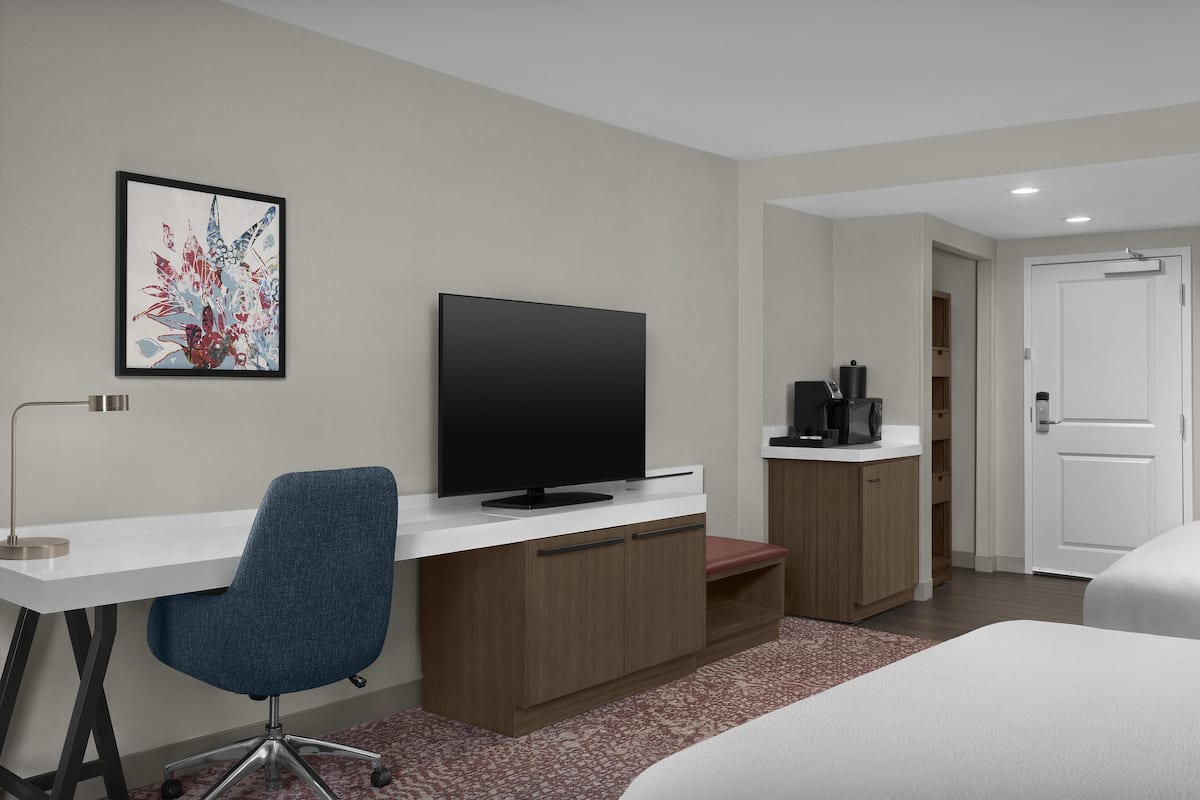 A hotel room with a flat-screen TV, a desk with a chair, a bed, a coffee machine, and a door.