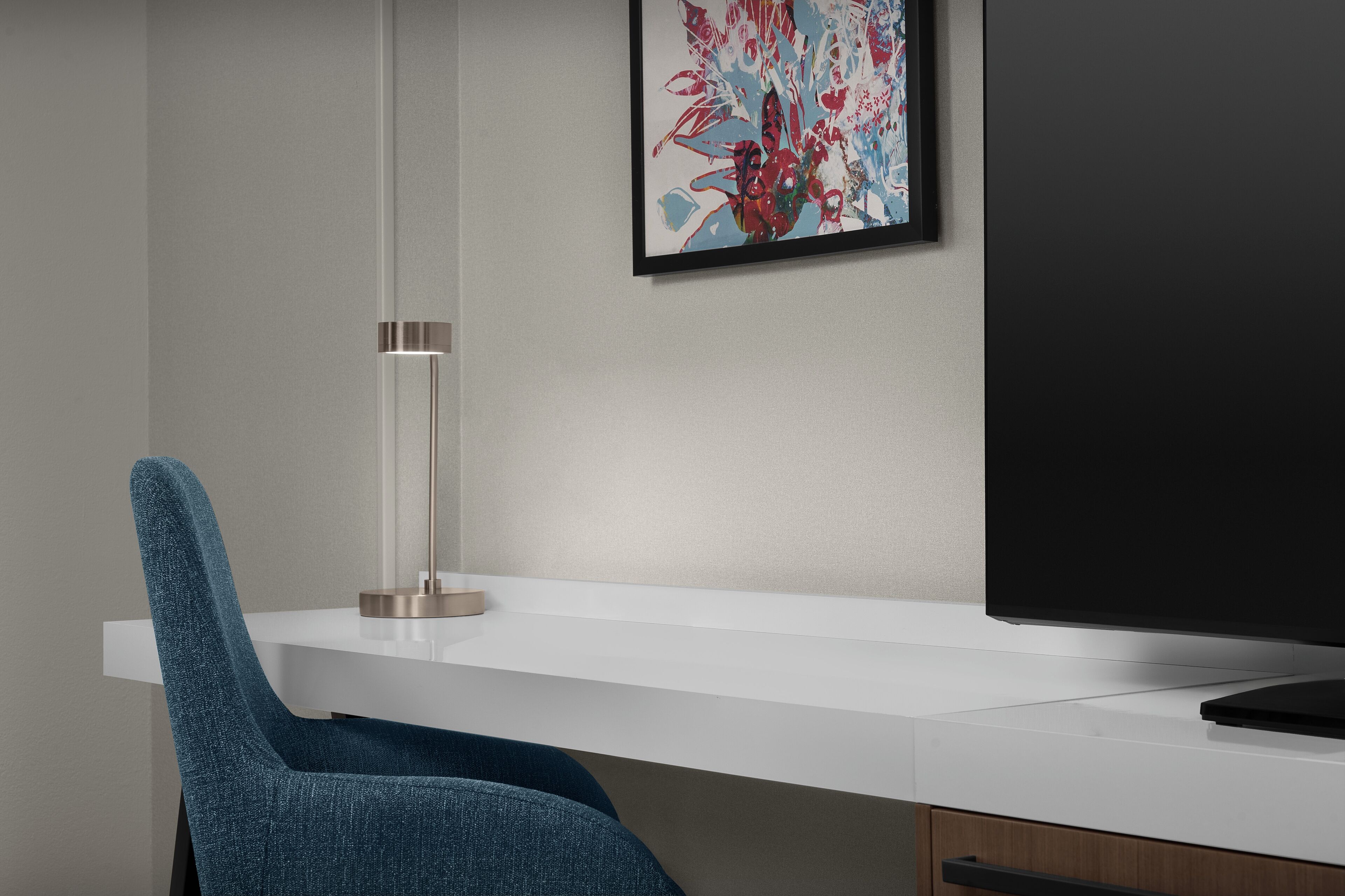 A modern office desk with a blue chair, a framed abstract painting, and a flat-screen TV.