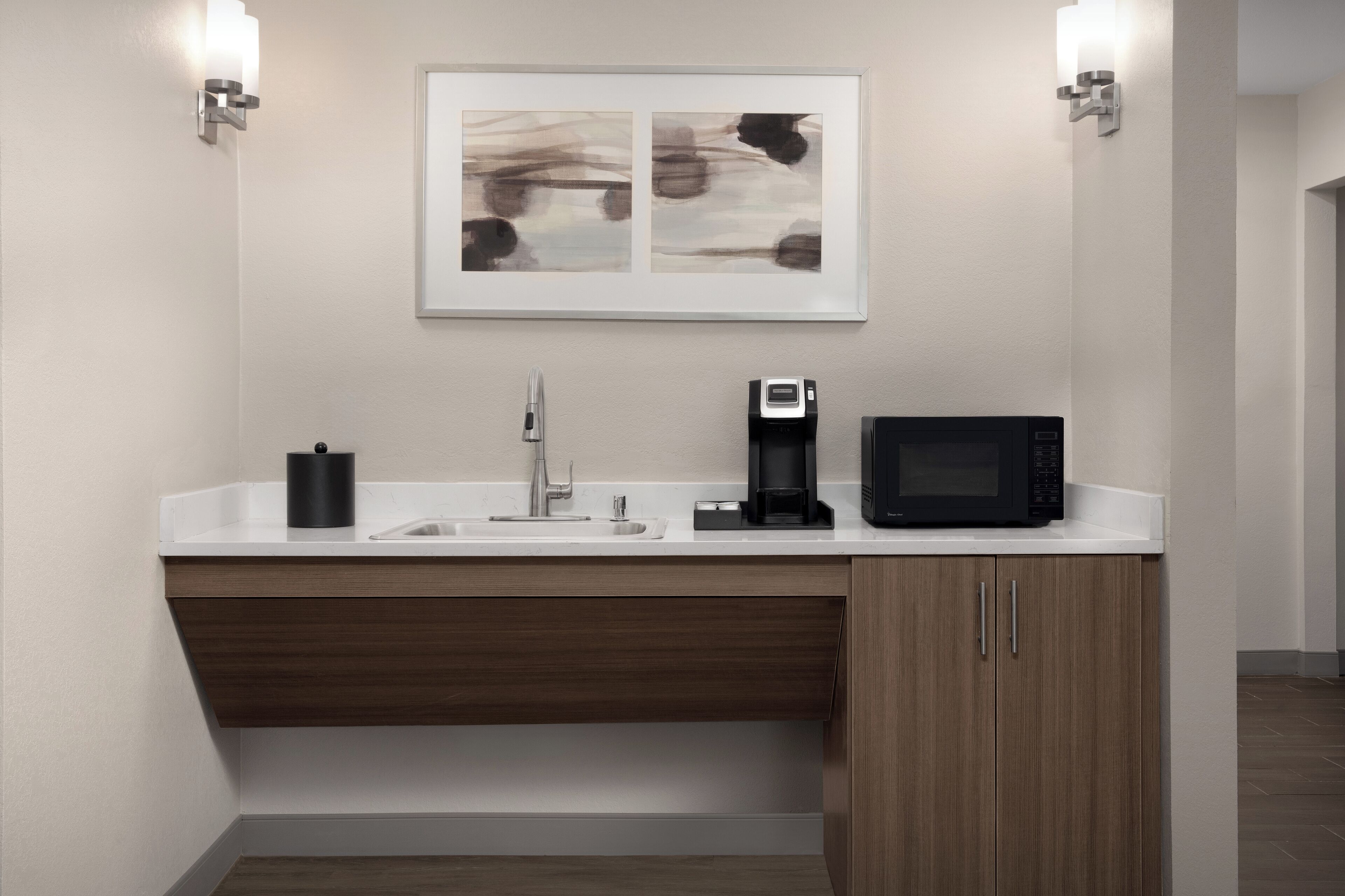 Un bany modern amb un lavabo blanc, una cafetera, un microones i un quadre abstracte encimbellat sobre el lavabo.