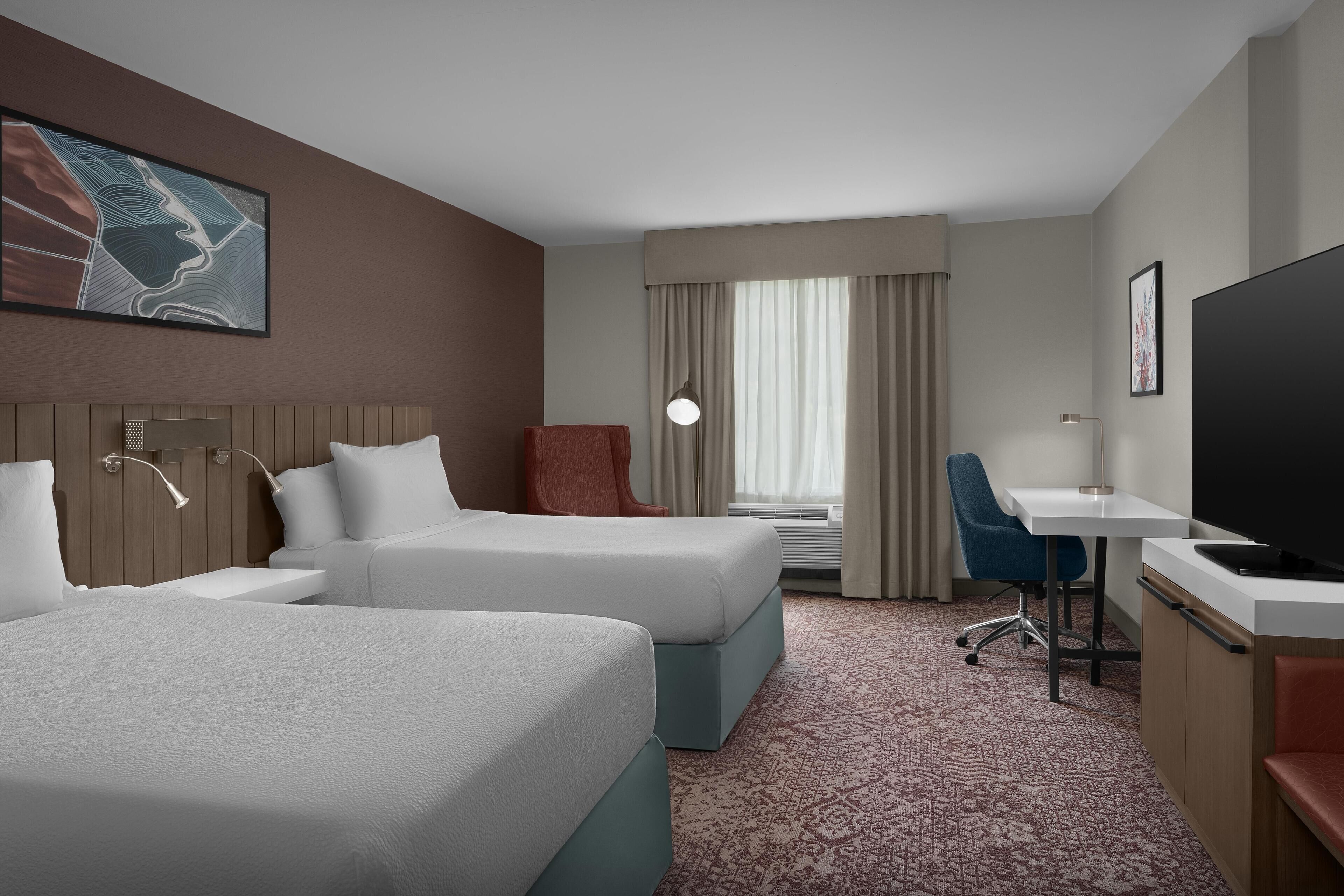 Habitación de hotel con dos camas, un escritorio, televisión y un amplio ventanal con cortinas.
