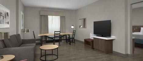 A modern hotel room with a grey sofa, a red ottoman, a wooden dining table with chairs, a TV on a wooden stand, and a bed with a blue headboard.