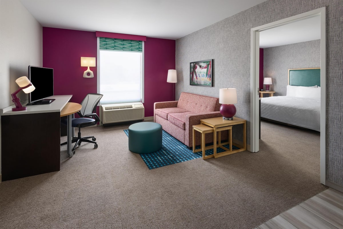 A hotel room with a desk, computer, chair, sofa, bedside tables, lamp, and a bed.