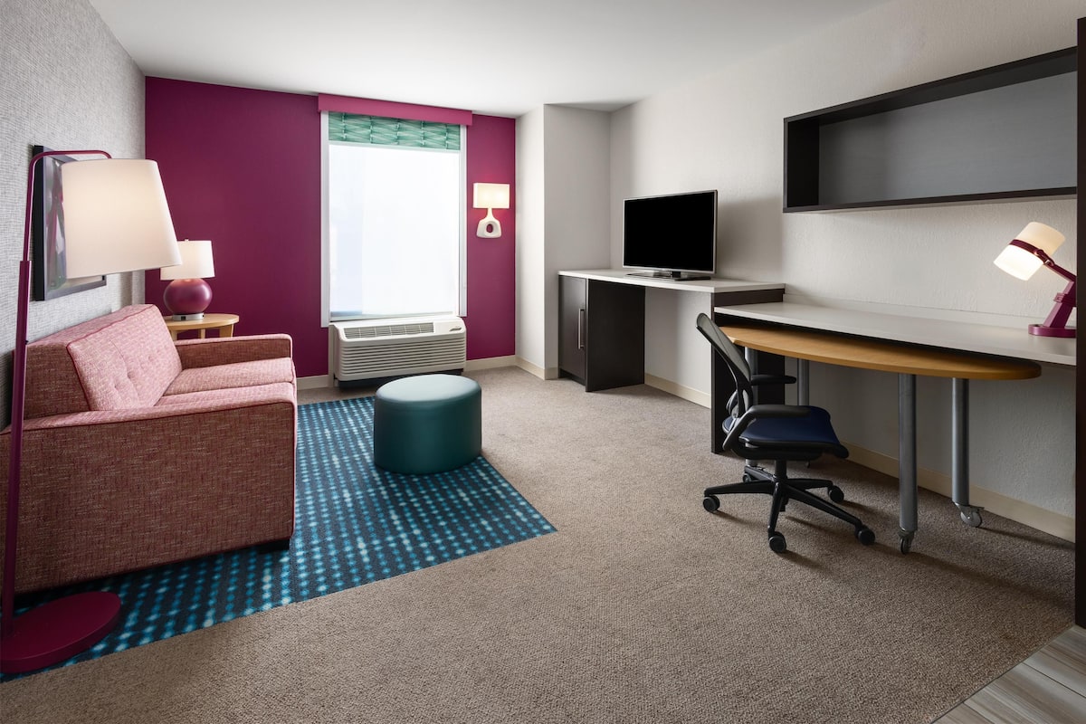 A modern hotel room with a pink sofa, a desk with a computer, a TV, and a small round ottoman.