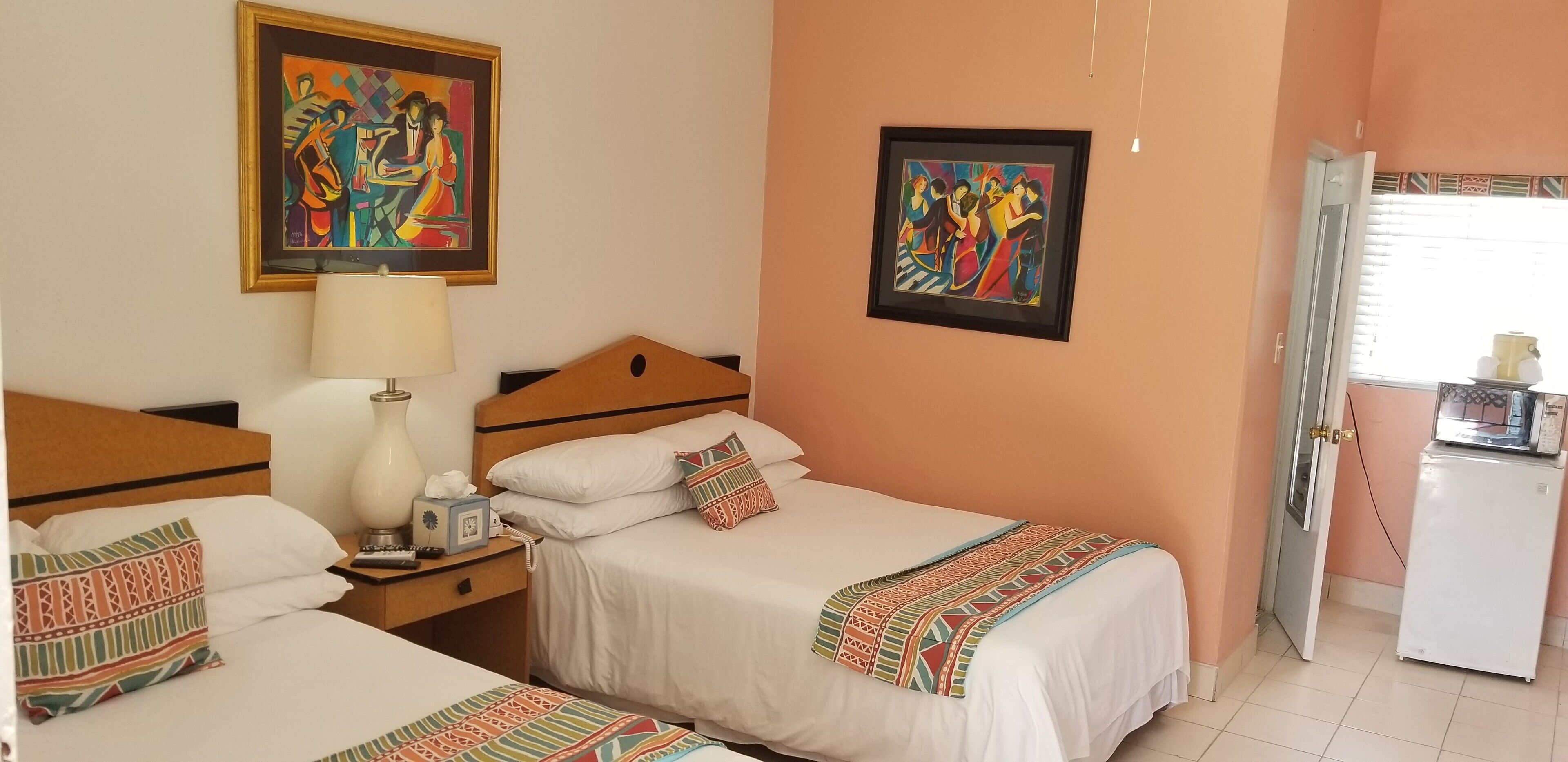A hotel room with two beds, a nightstand with a lamp, a small table with a clock, and two framed paintings on the wall.