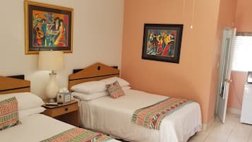 A hotel room with two beds, a nightstand with a lamp, a small table with a clock, and two framed paintings on the wall.